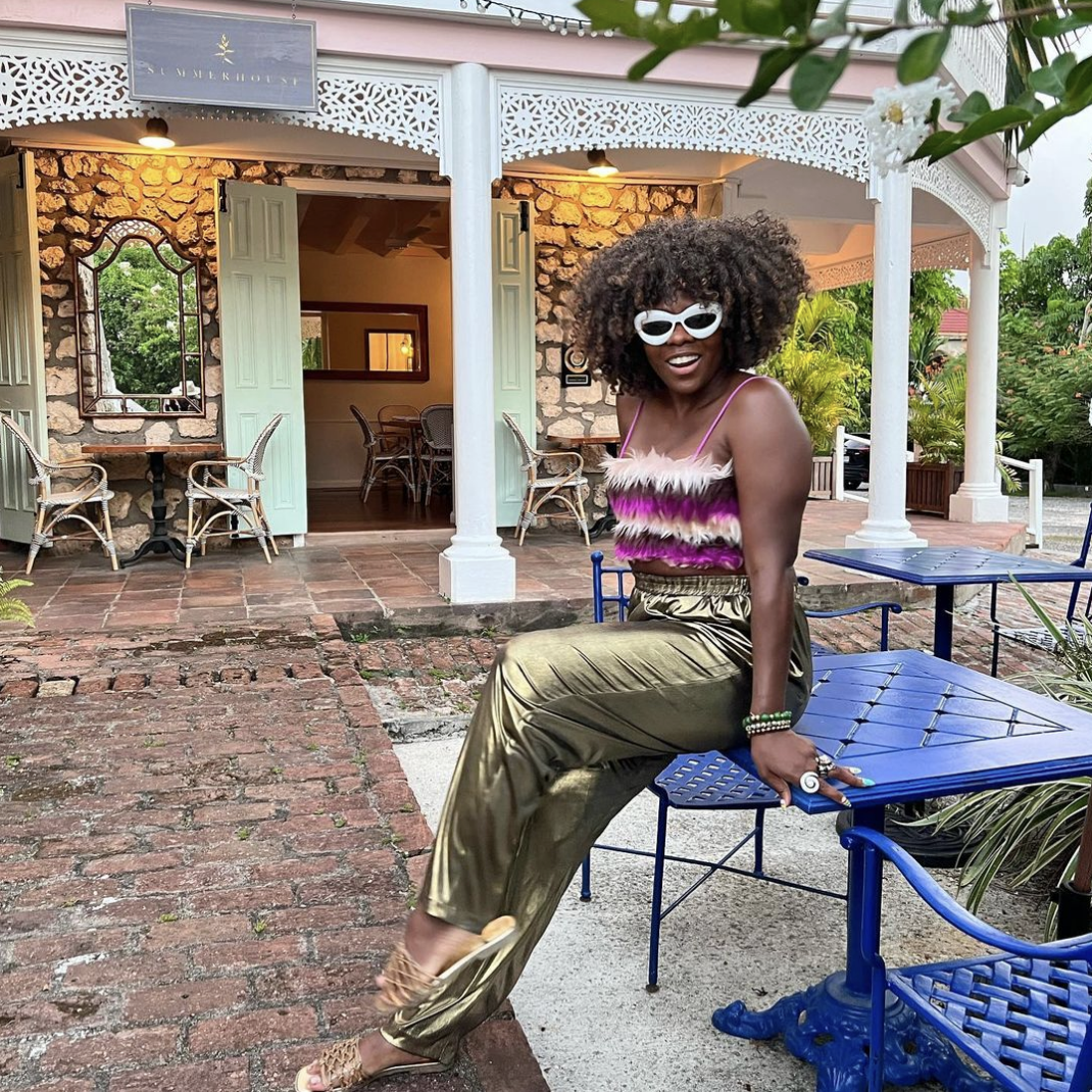 Patrice in a fur cami top and satin pants seated at an outdoor cafe with a historic building in the background