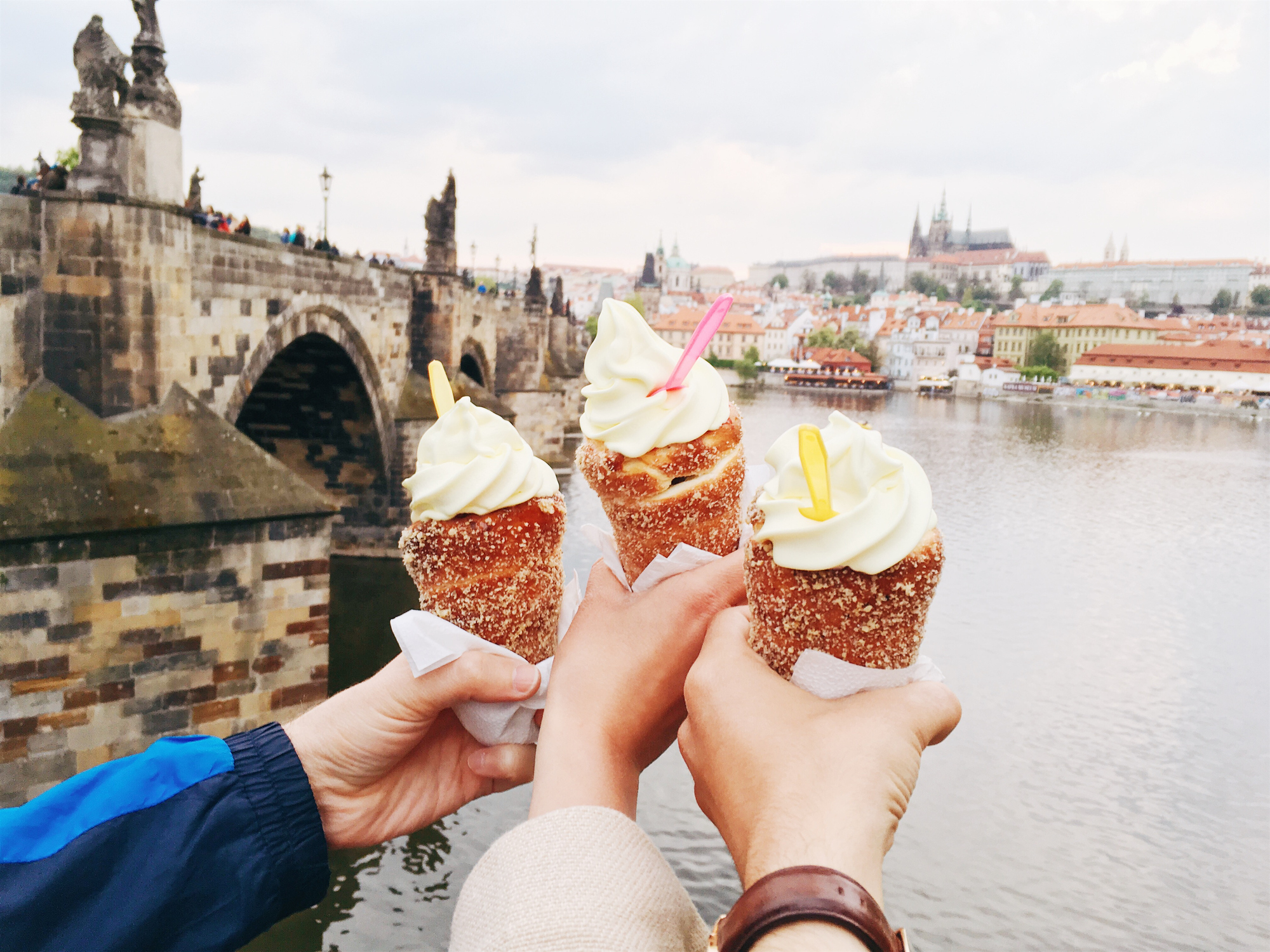 Three hands holding ice cream cones in front of a historic bridge