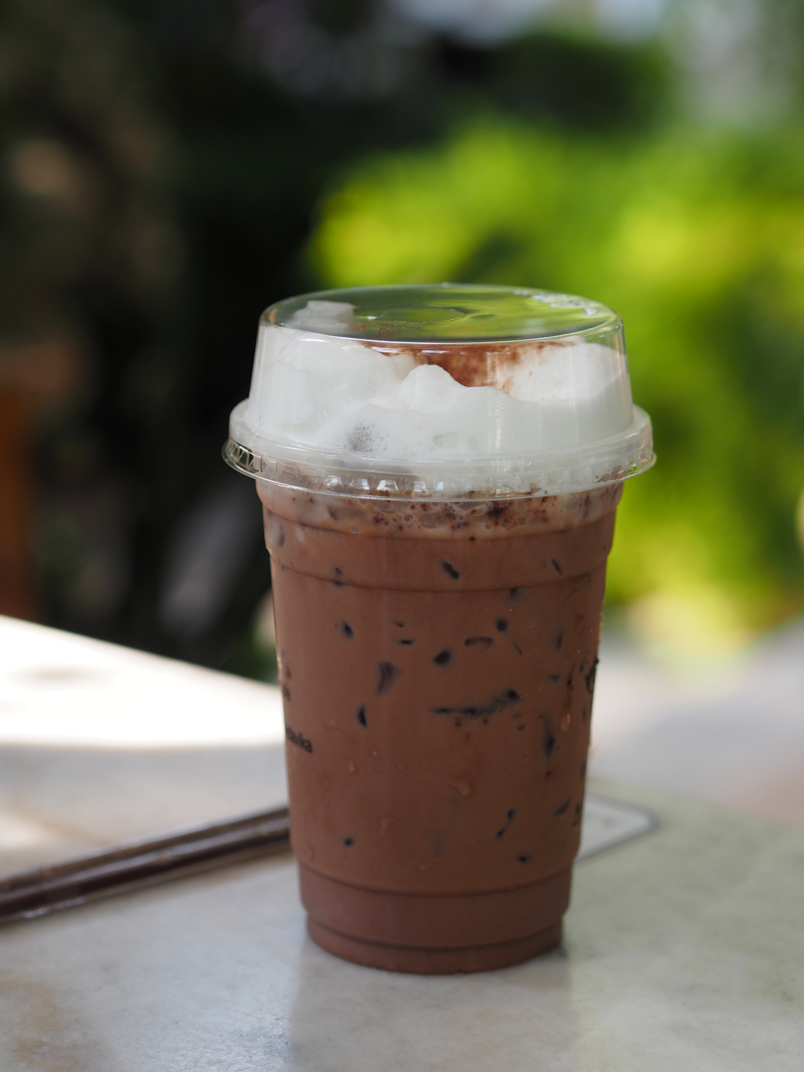 Iced chocolate drink with whipped cream in a clear cup, outdoors on a table with a blurred natural background