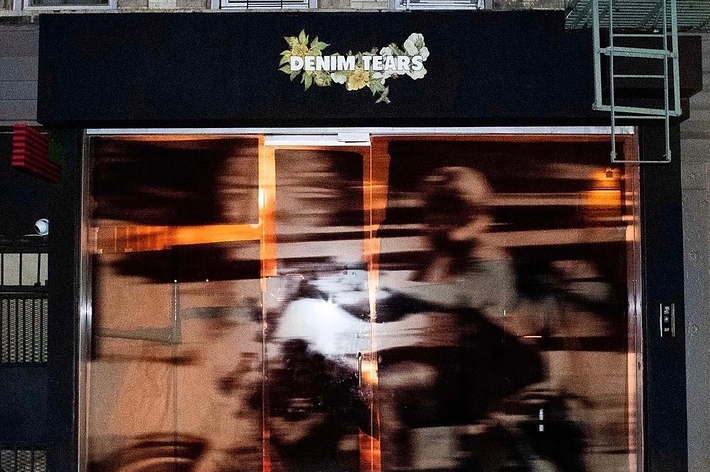An obscured view of a storefront with the sign 'Denim Tears' above; reflections and shadows create an abstract effect