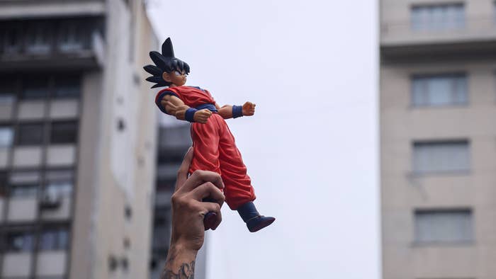 Hand holding an action figure of Goku from Dragon Ball in the air with buildings as backdrop