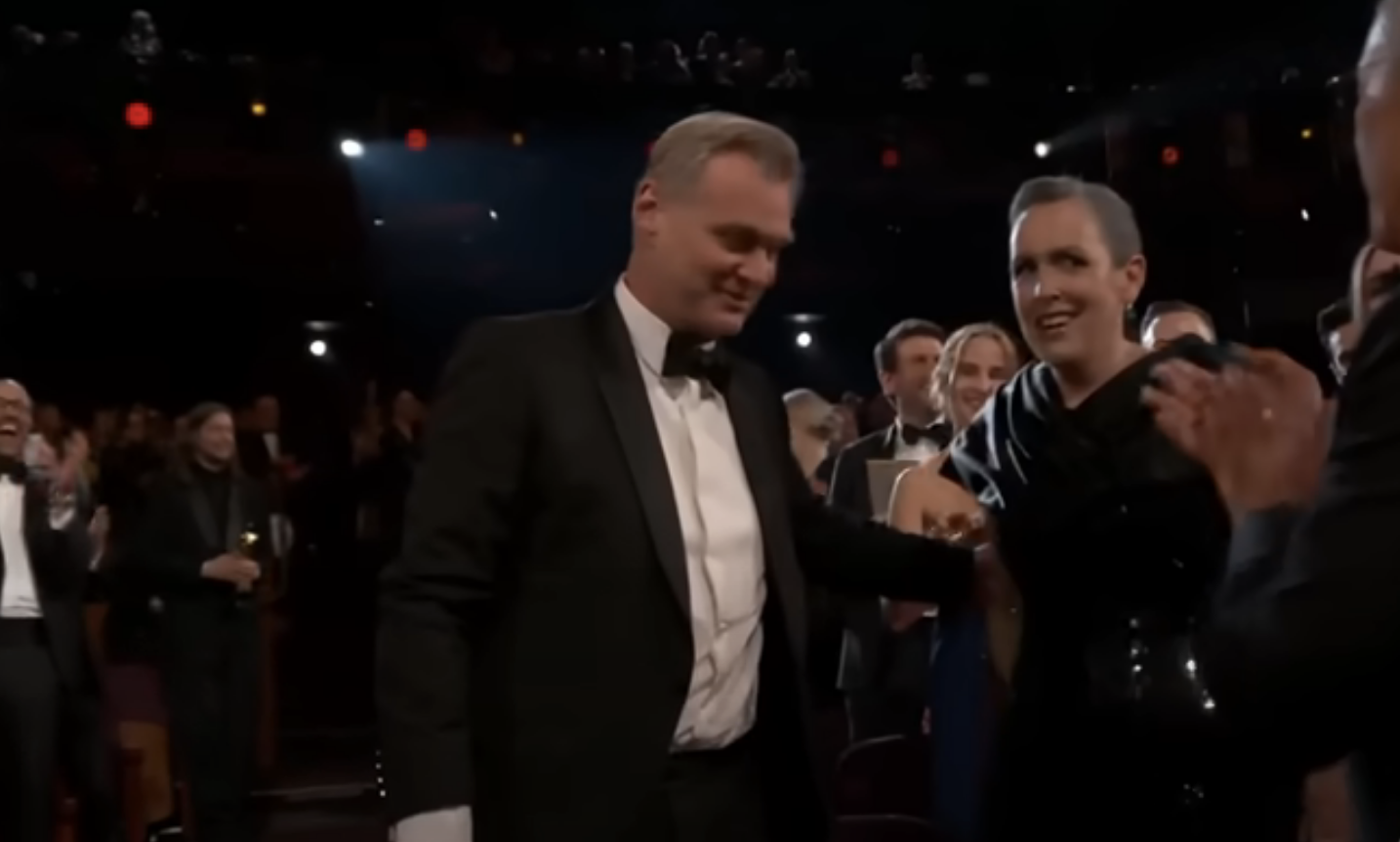 Christopher Nolan and Emma embracing in the audience, surrounded by applauding attendees