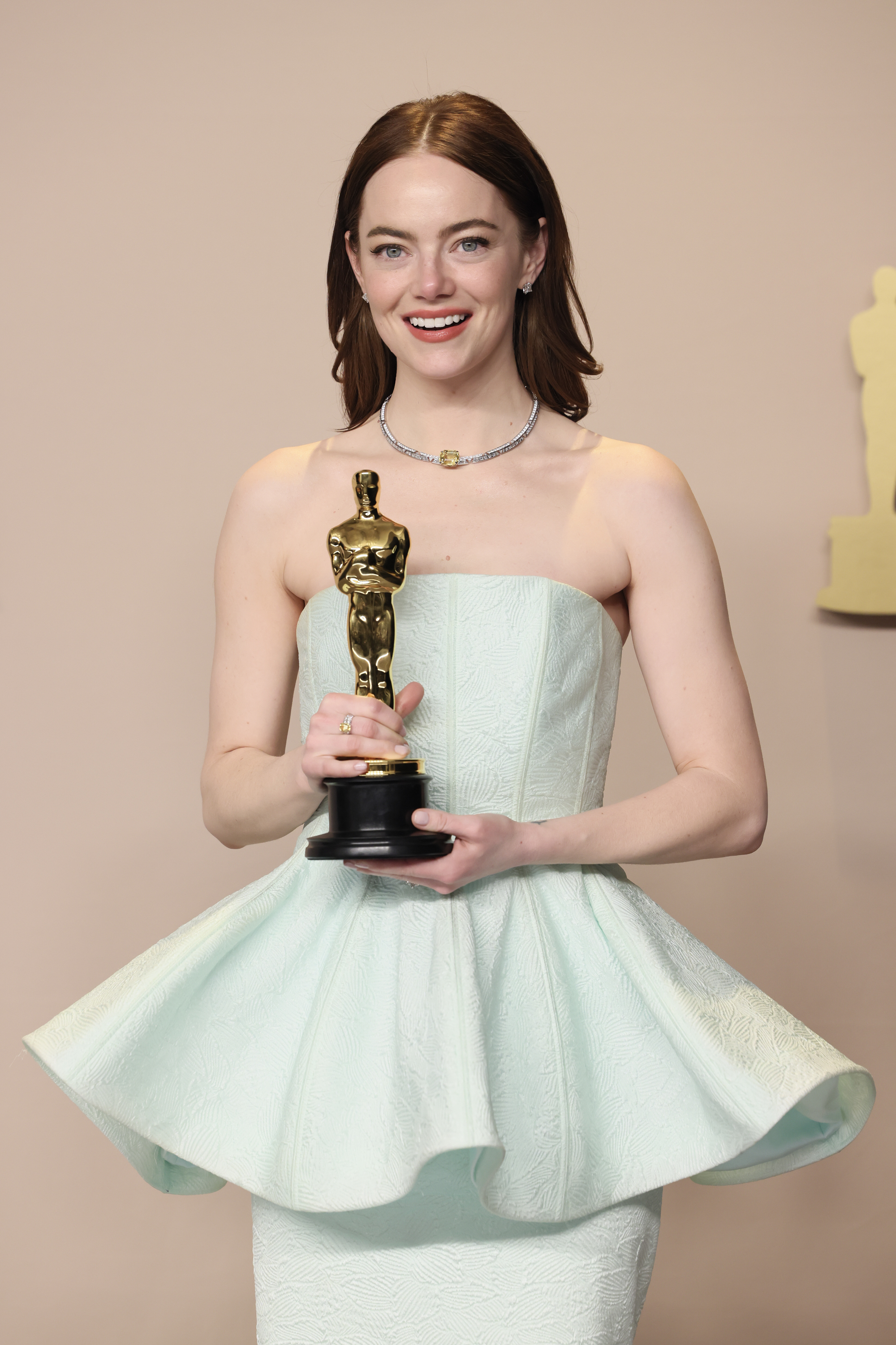 Emma Stone posing with an Oscar, wearing a strapless gown with a peplum waist