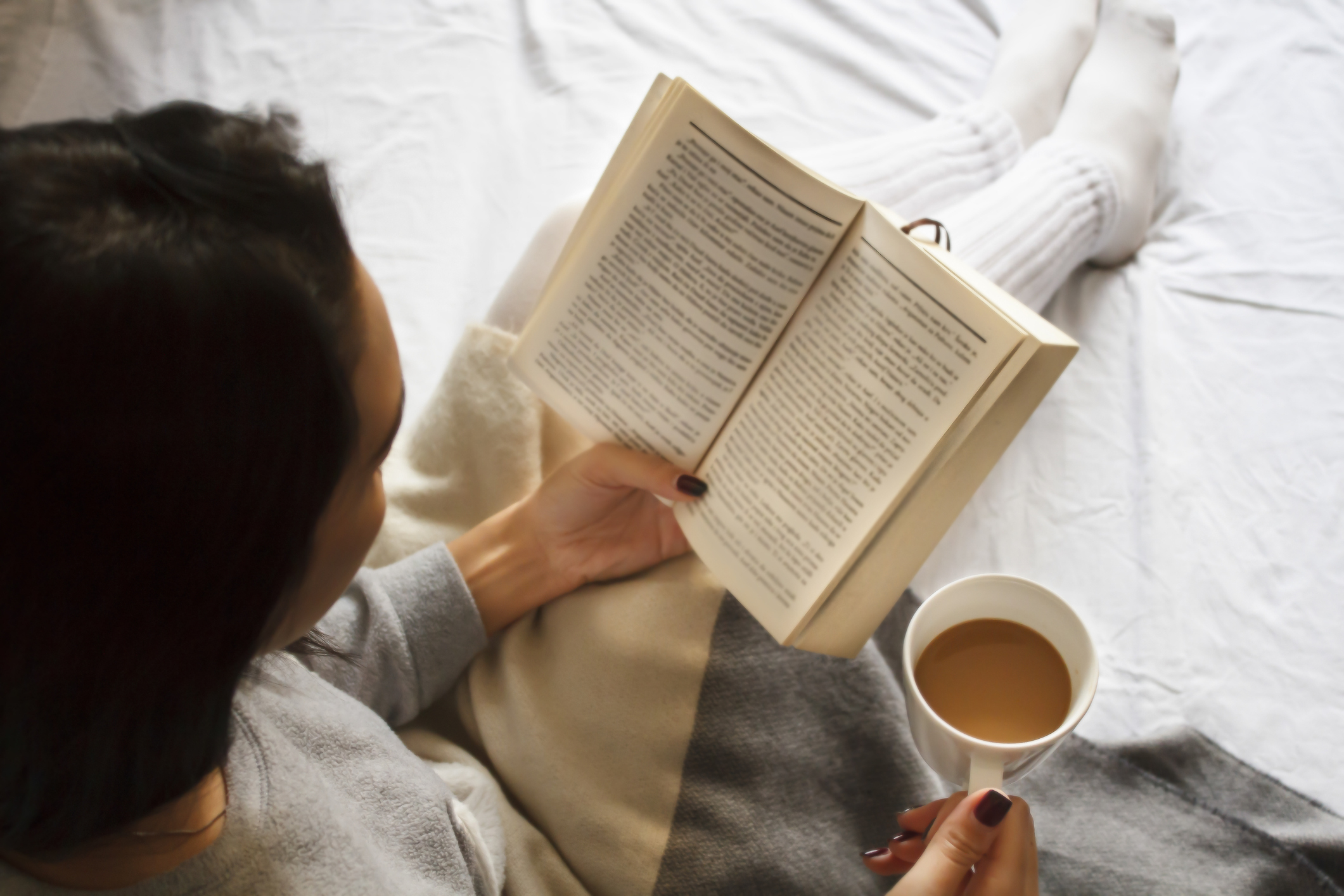 Person reading a book and holding a cup of coffee while sitting on a bed