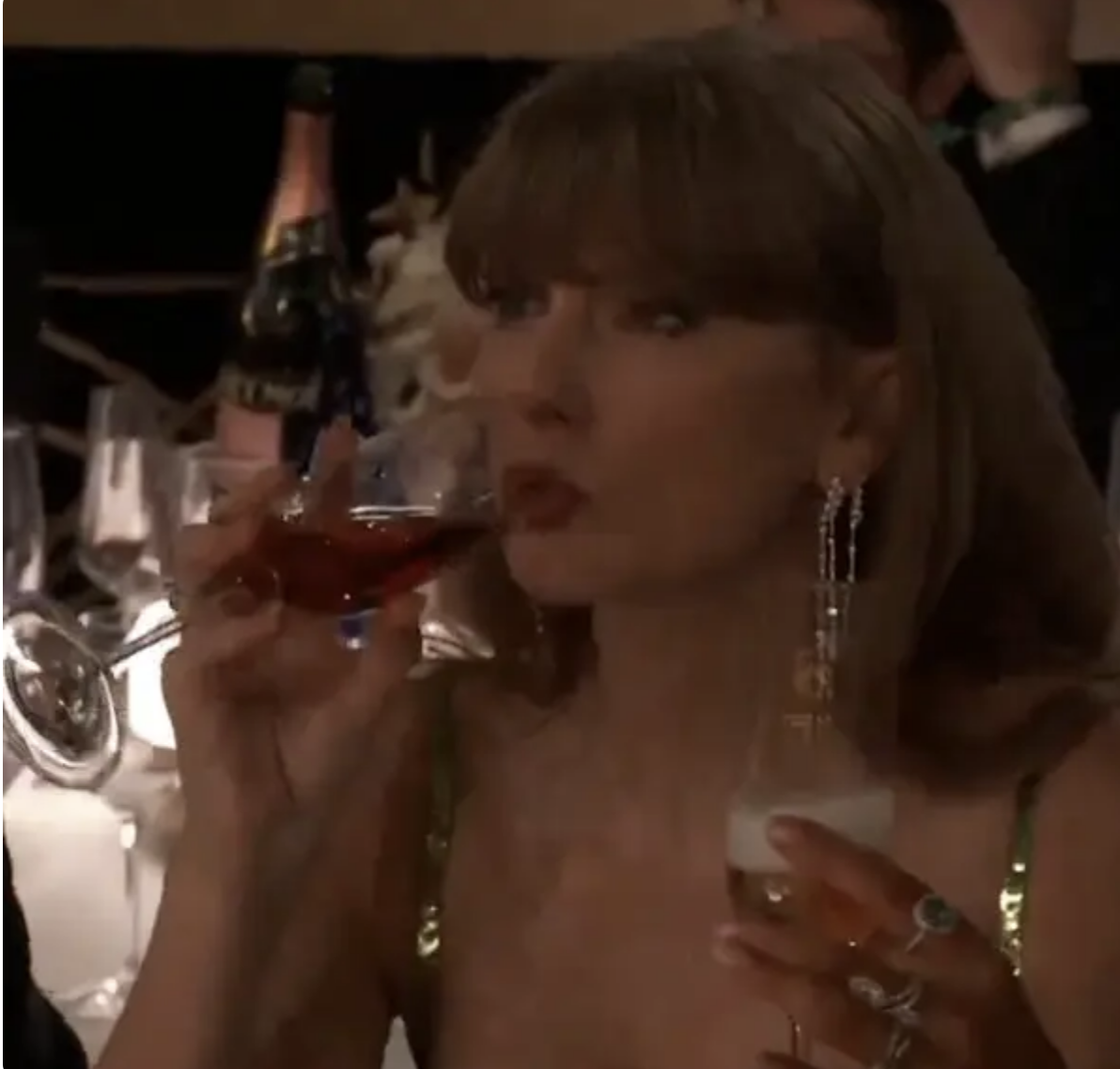 Taylor Swift sipping a drink at an event, wearing sparkling earrings and a sequined dress