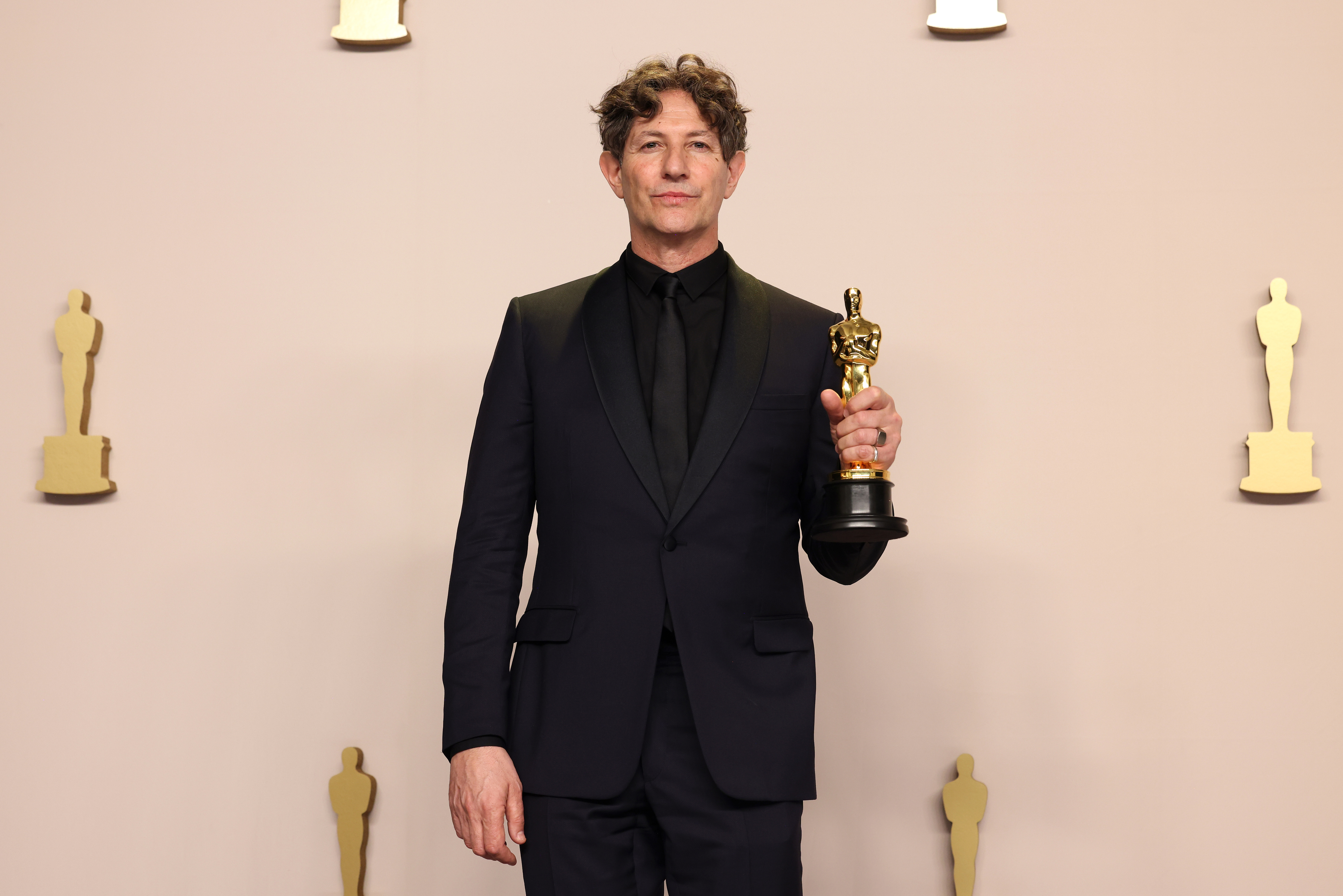 Jonathan holding an Oscar trophy, dressed in a black suit and tie, standing in front of statue silhouettes