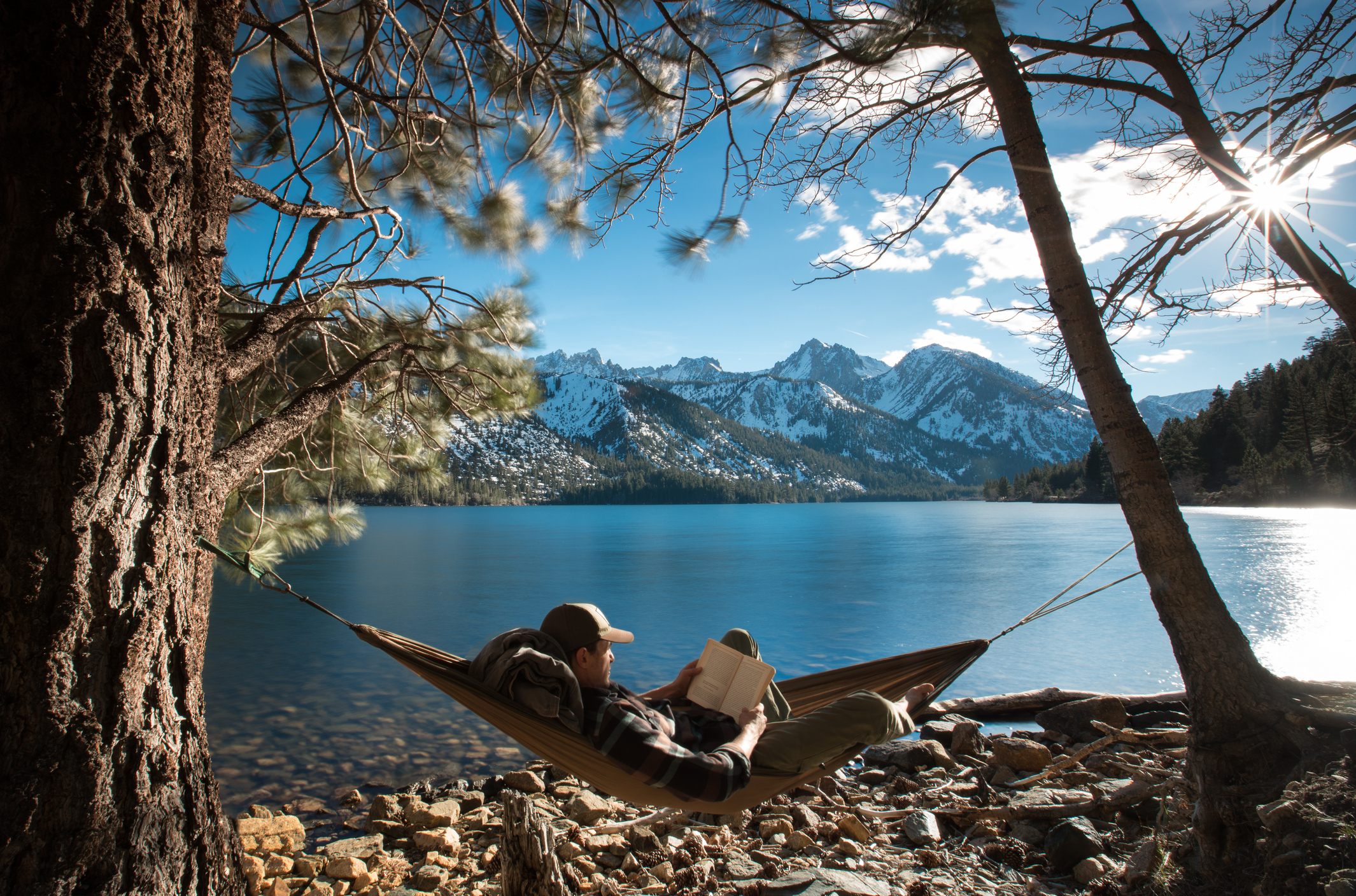 Person in a hammock by a lake, reading a book, with mountains in the background