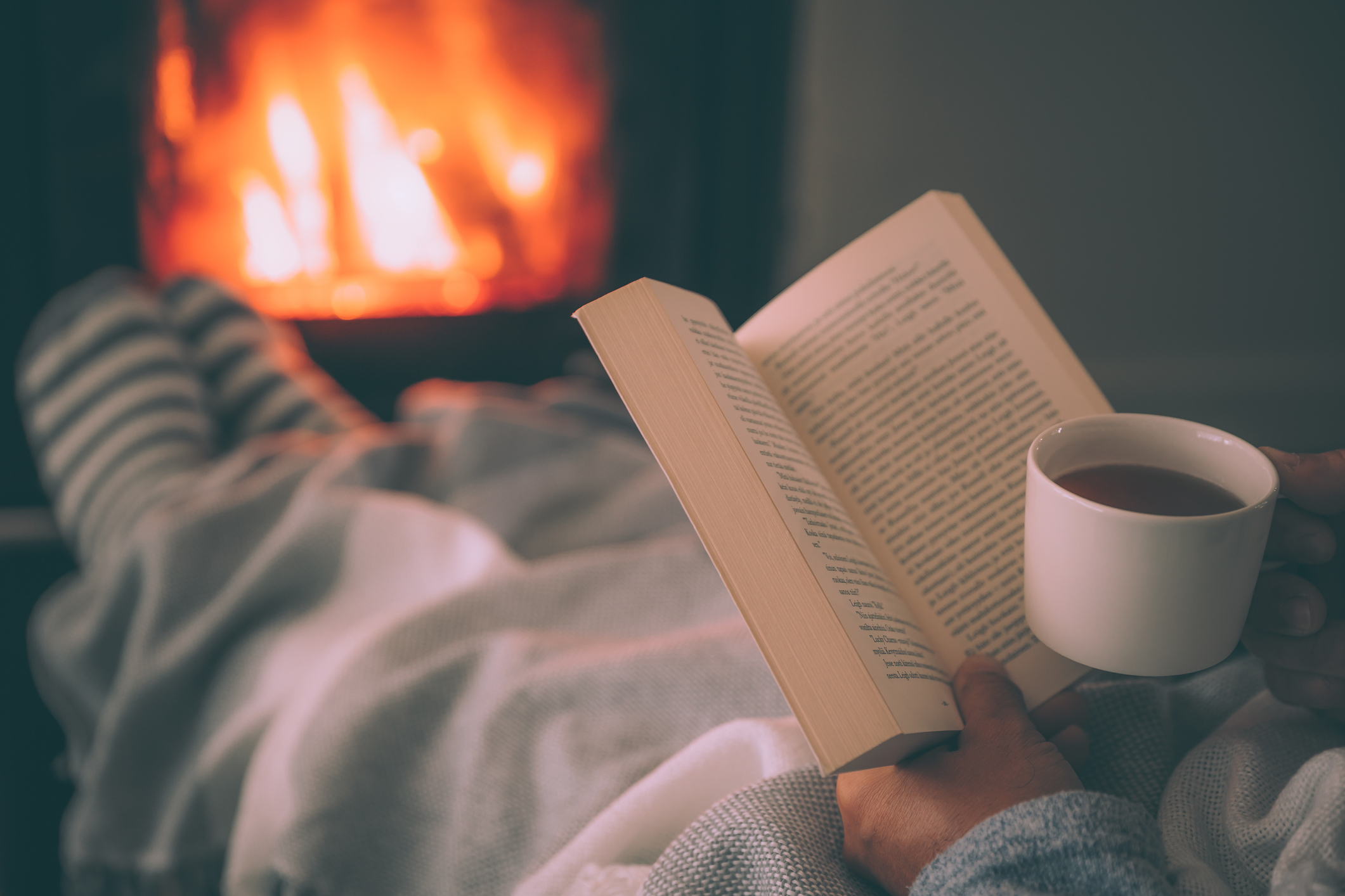 Person relaxing with a book and a cup of tea by a fireplace