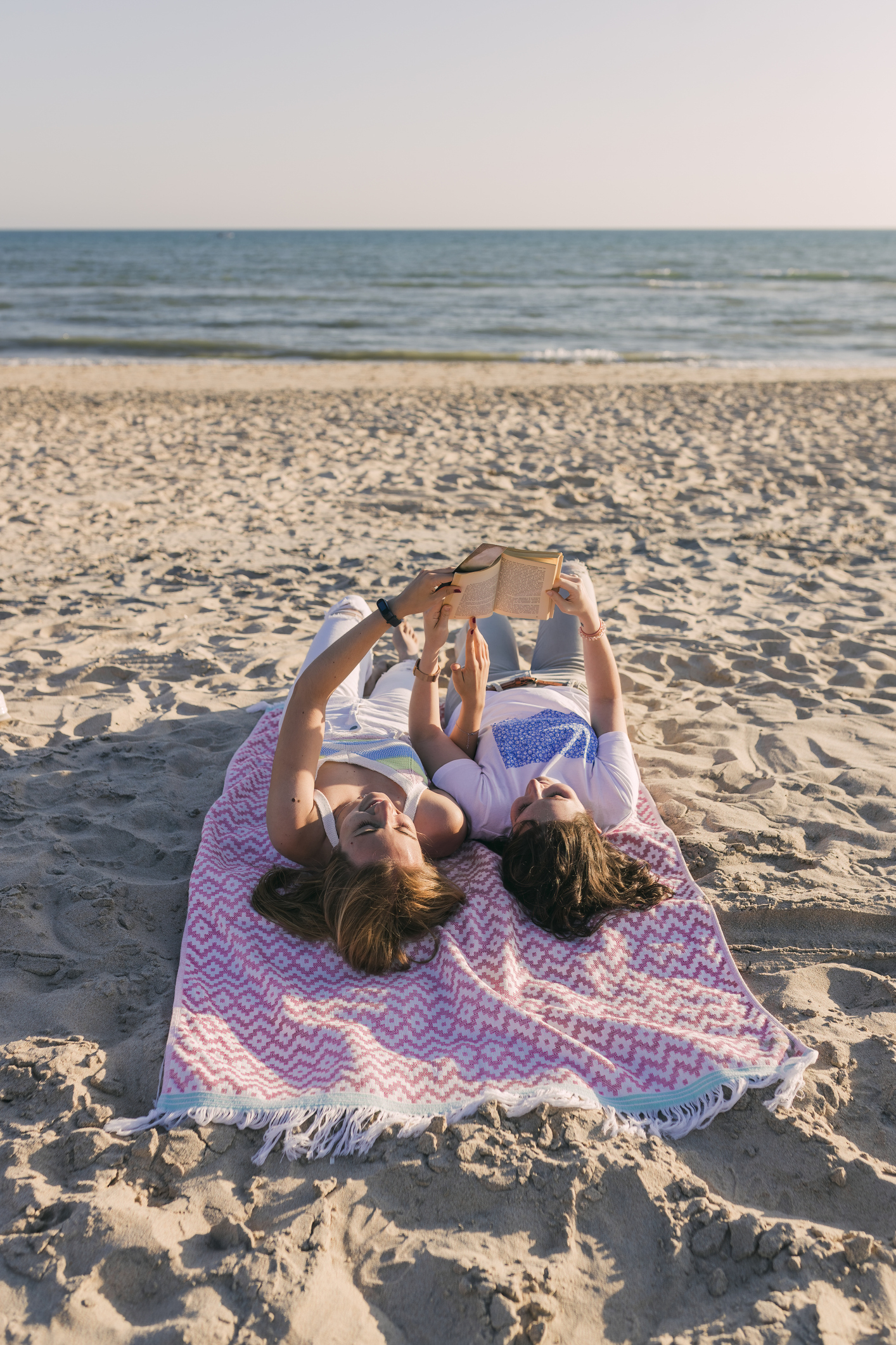 Two individuals lying on a beach towel, one holding a book over both their heads