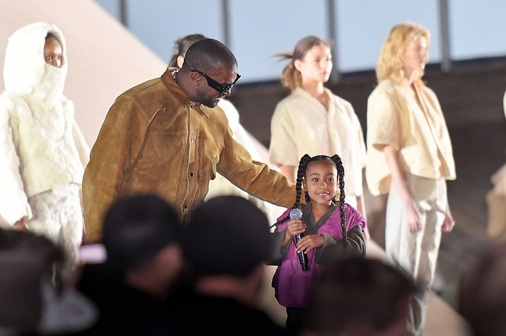 Man in a suede outfit supports a young girl holding a microphone on stage, with choir members in the background