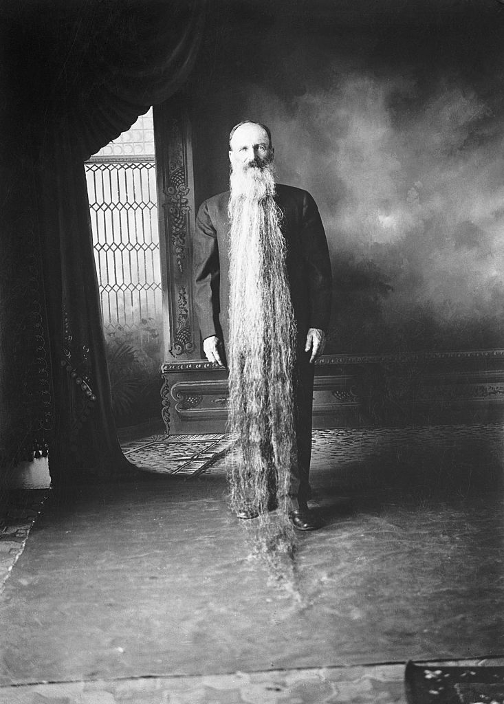 Man with an extremely long beard standing in a room, next to a draped curtain and ornate furniture