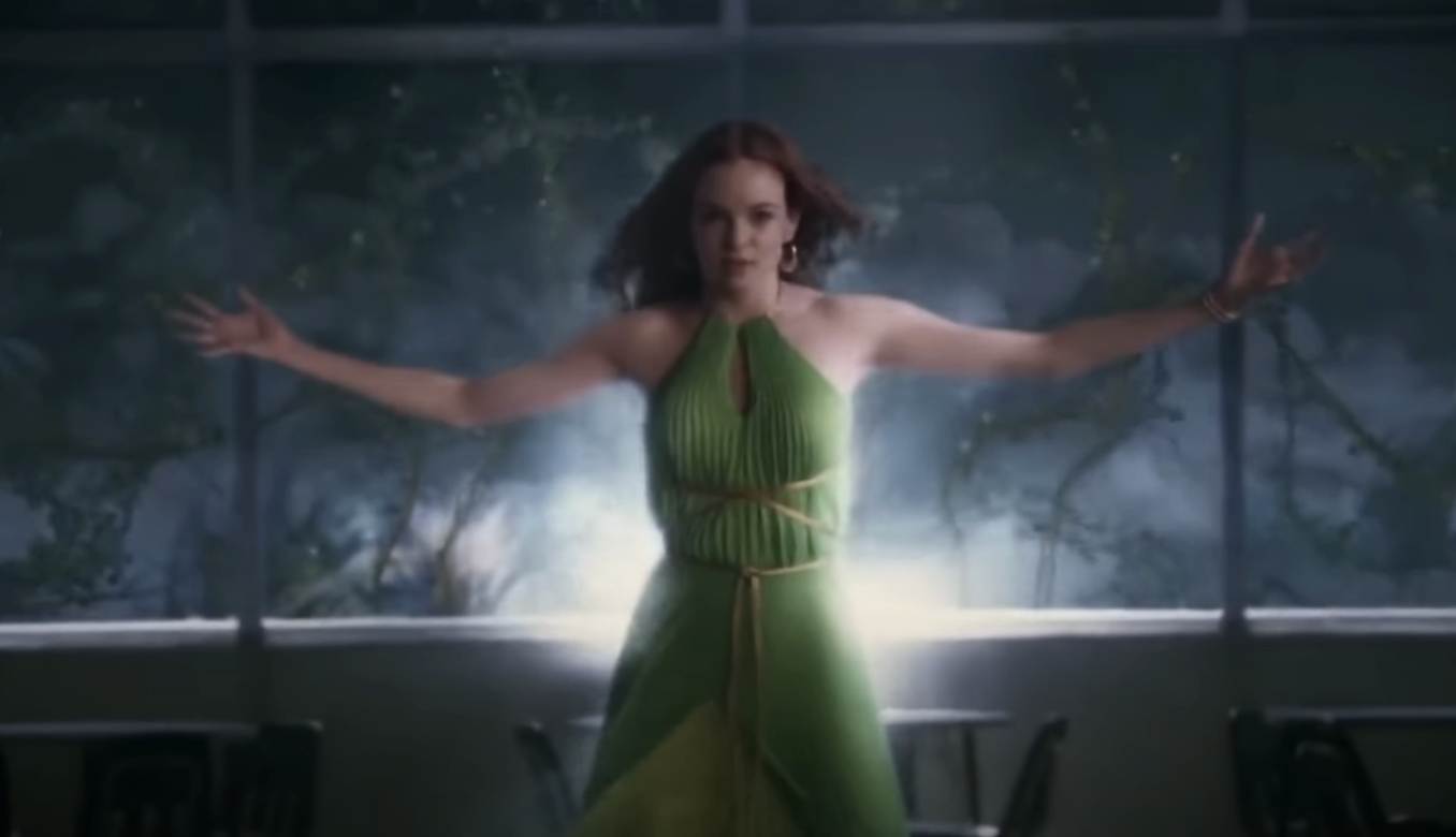 Woman in a green pleated dress with outstretched arms, standing in a room with windows in the background