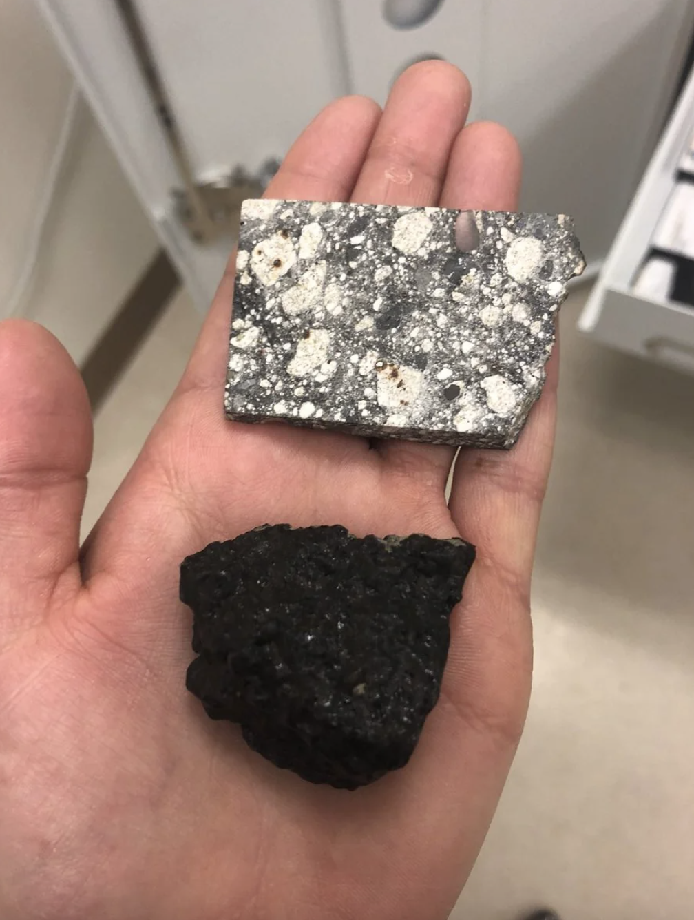 A person&#x27;s hand holding a speckled square mineral sample above a rough, irregular-shaped black sample