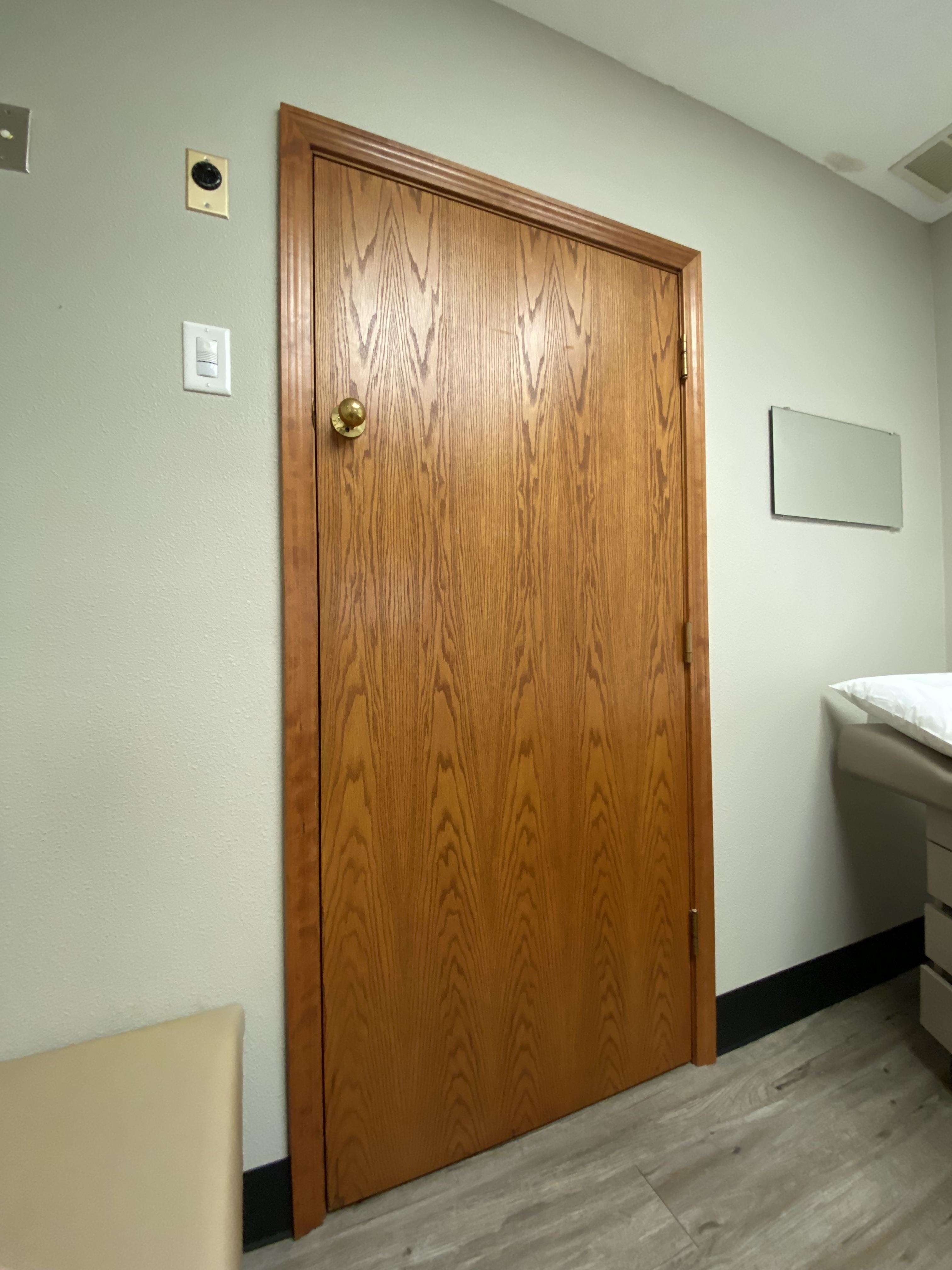 A closed wooden door in a room with a whiteboard and examination bed on the right side, with the knob more than halfway up the door