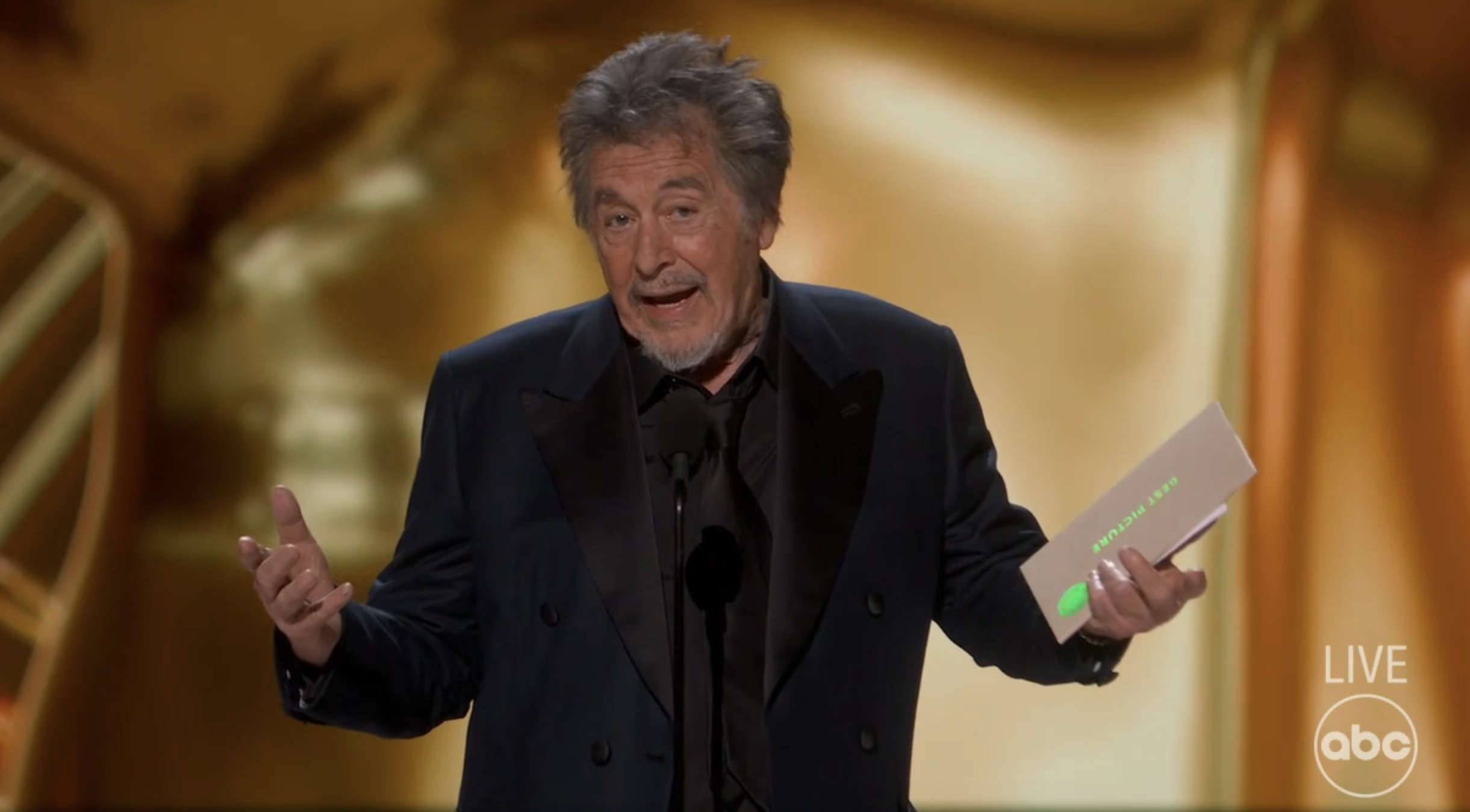 Al Pacino speaks on stage at an awards show, holding an envelope and gesturing with his hand