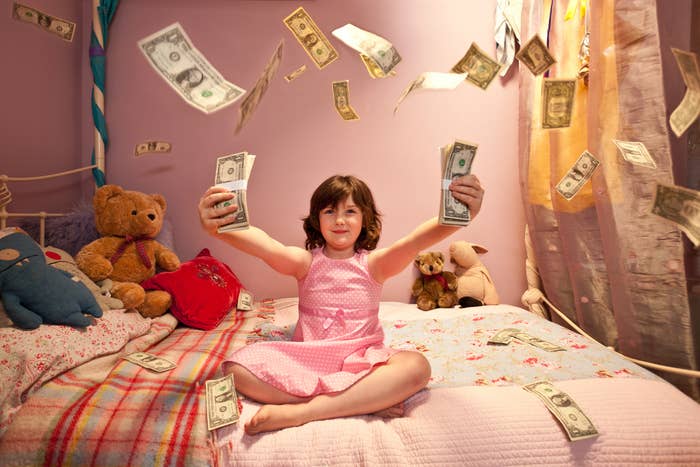 A child in a pink dress smiles, holding up a dollar bill with money scattered around in a bedroom