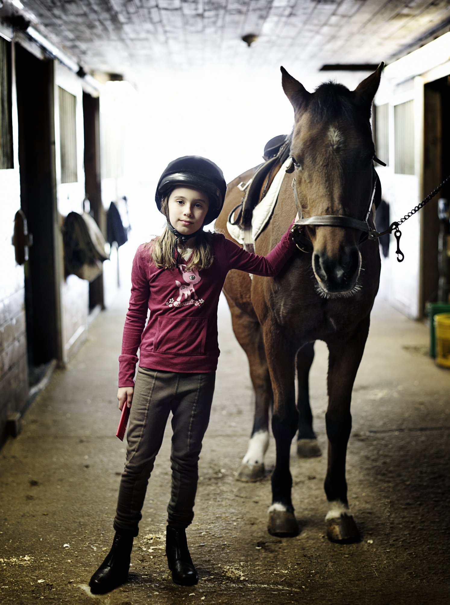 Child in riding helmet stands next to a horse in a stable