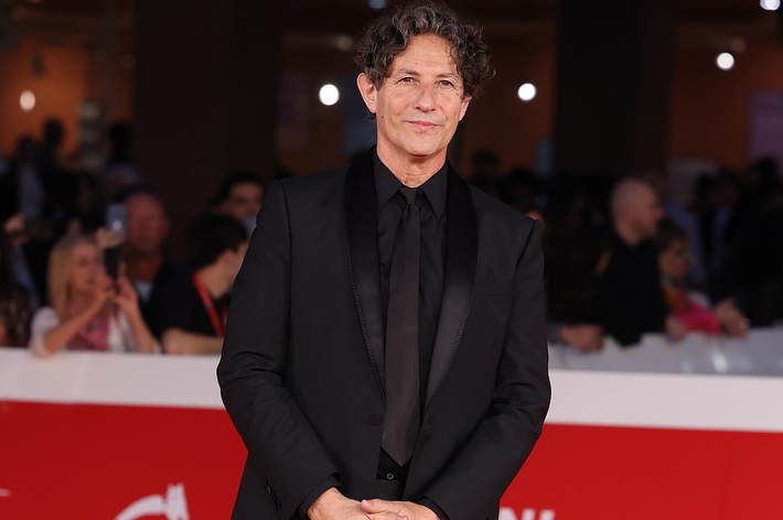 Man in a black suit smiles at a red carpet event, hands clasped in front