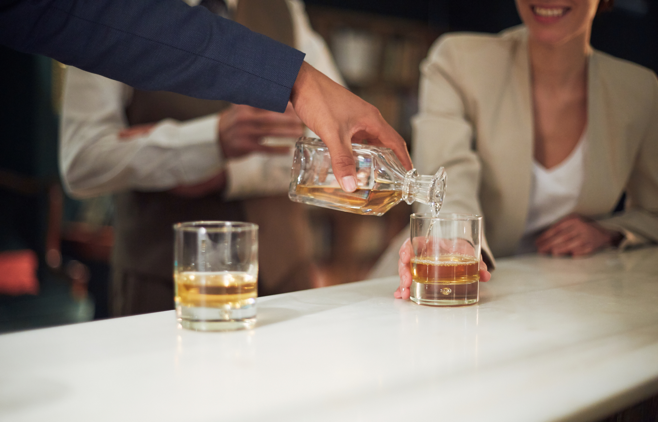 Person pouring whiskey into glasses on a bar counter with smiling people in background