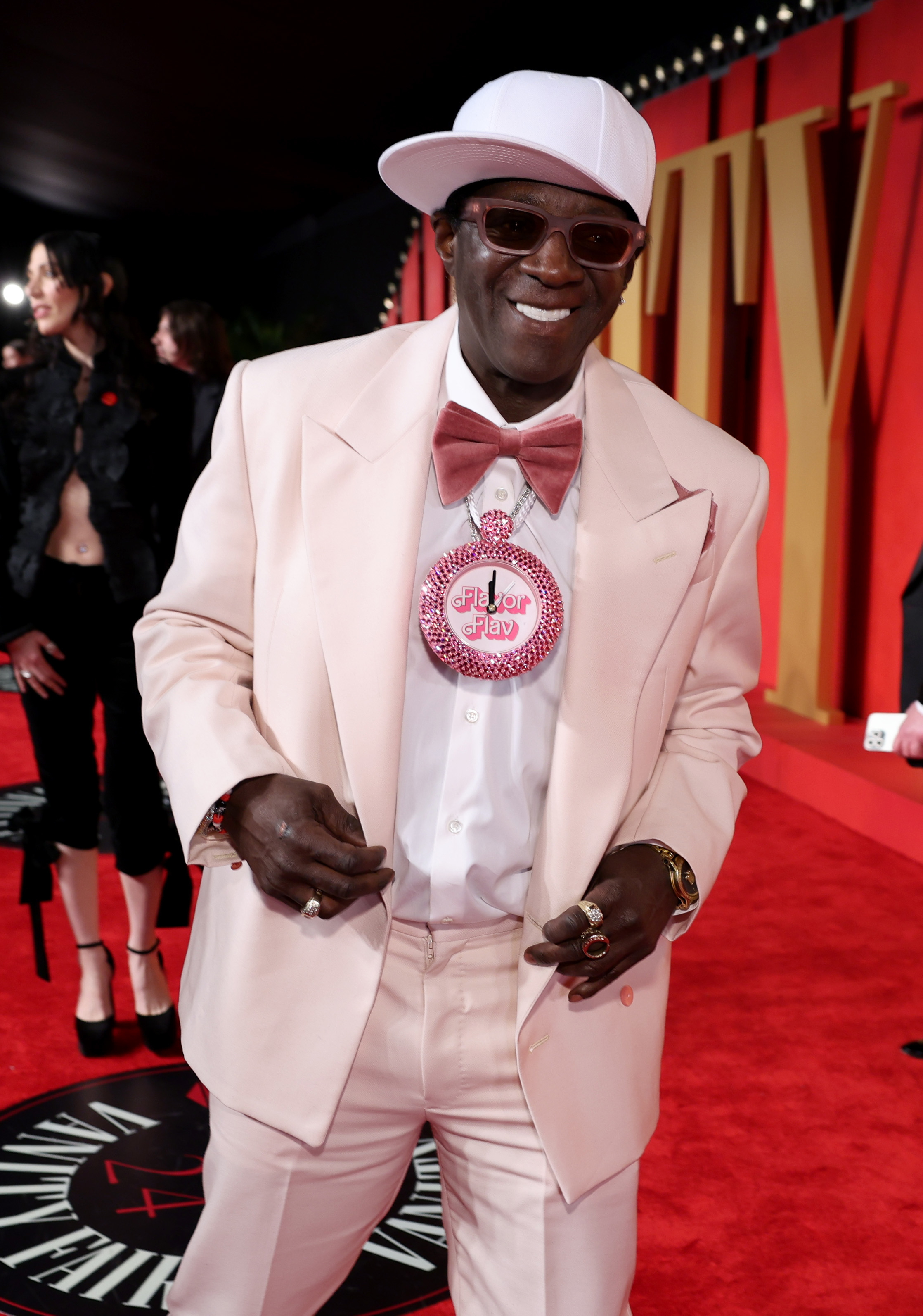 Flavor Flav in a light suit with a large clock necklace on a red carpet