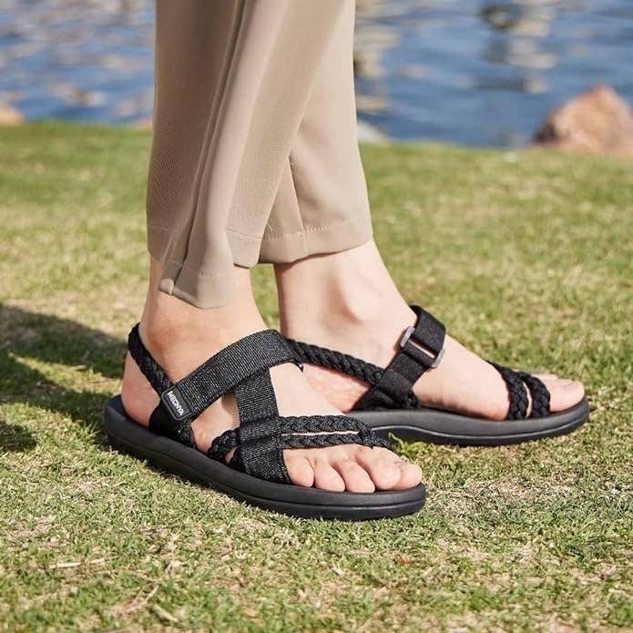 Person wearing black strappy sandals, shown from the shins down, standing on grass by water