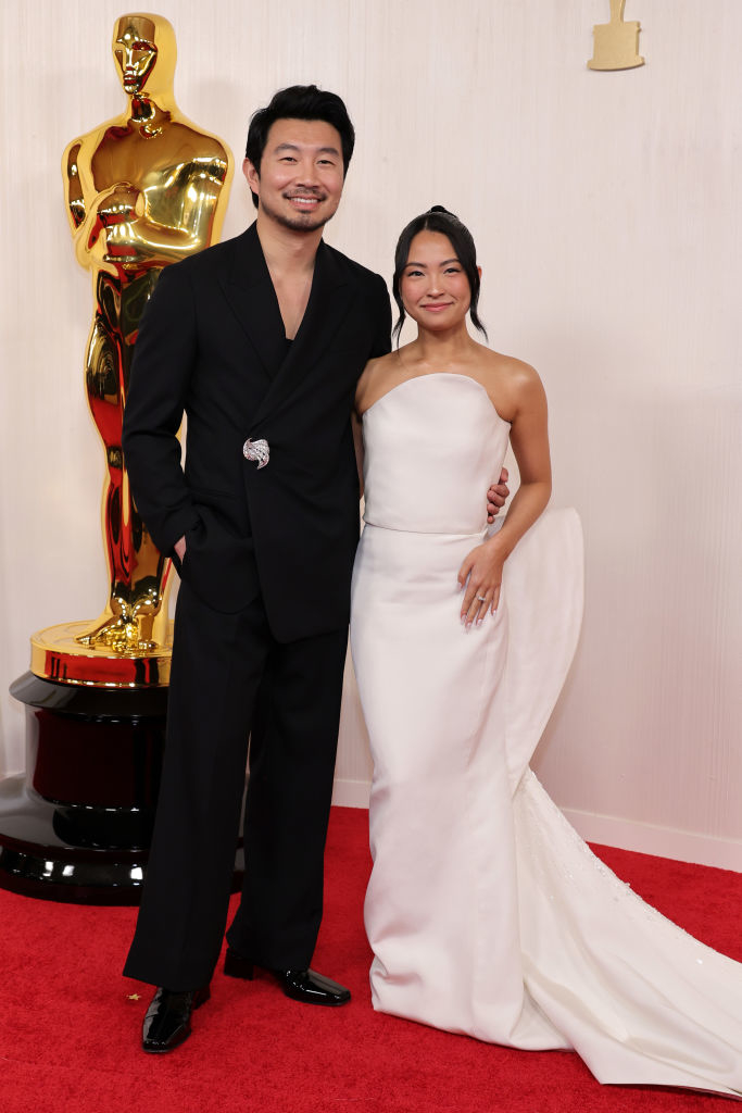 Two people posing on the red carpet; the person on the left in a black suit with emblem, and on the right in a white off-shoulder gown with a train