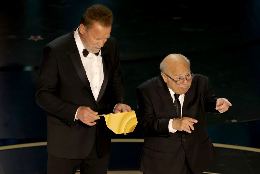 Two men in formal attire presenting on stage, one standing with a paper, the other gesturing