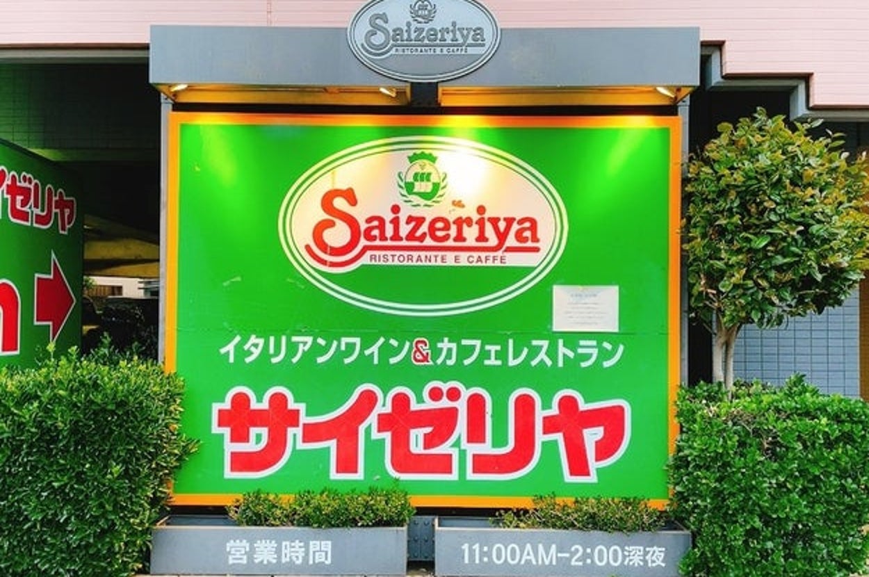 Front view of a Saizeriya restaurant entrance with logo and Japanese text, flanked by green bushes
