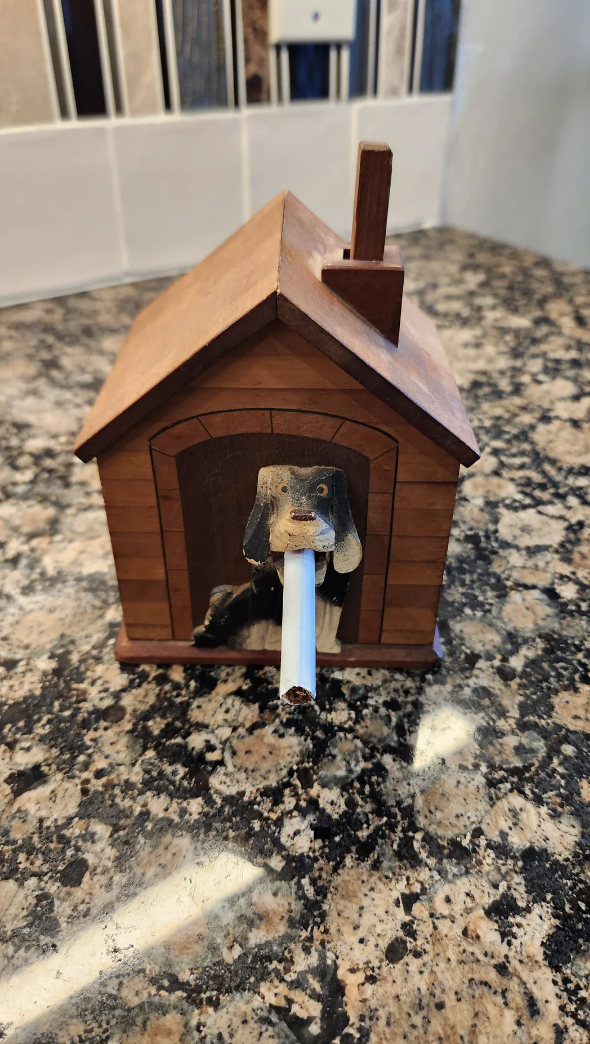 A small wooden dog&#x27;s face peeks out of a small wooden house, holding a tiny cigarette in its mouth
