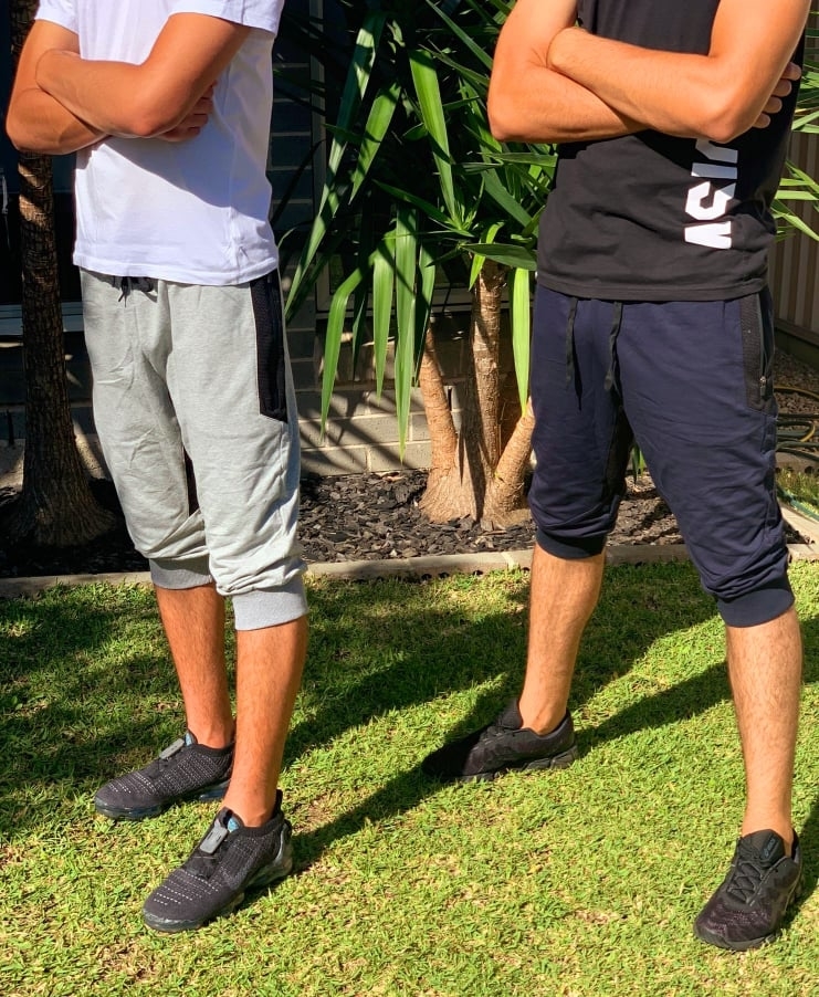 Two individuals standing side by side with arms crossed, wearing casual shorts and sneakers, outdoors near foliage