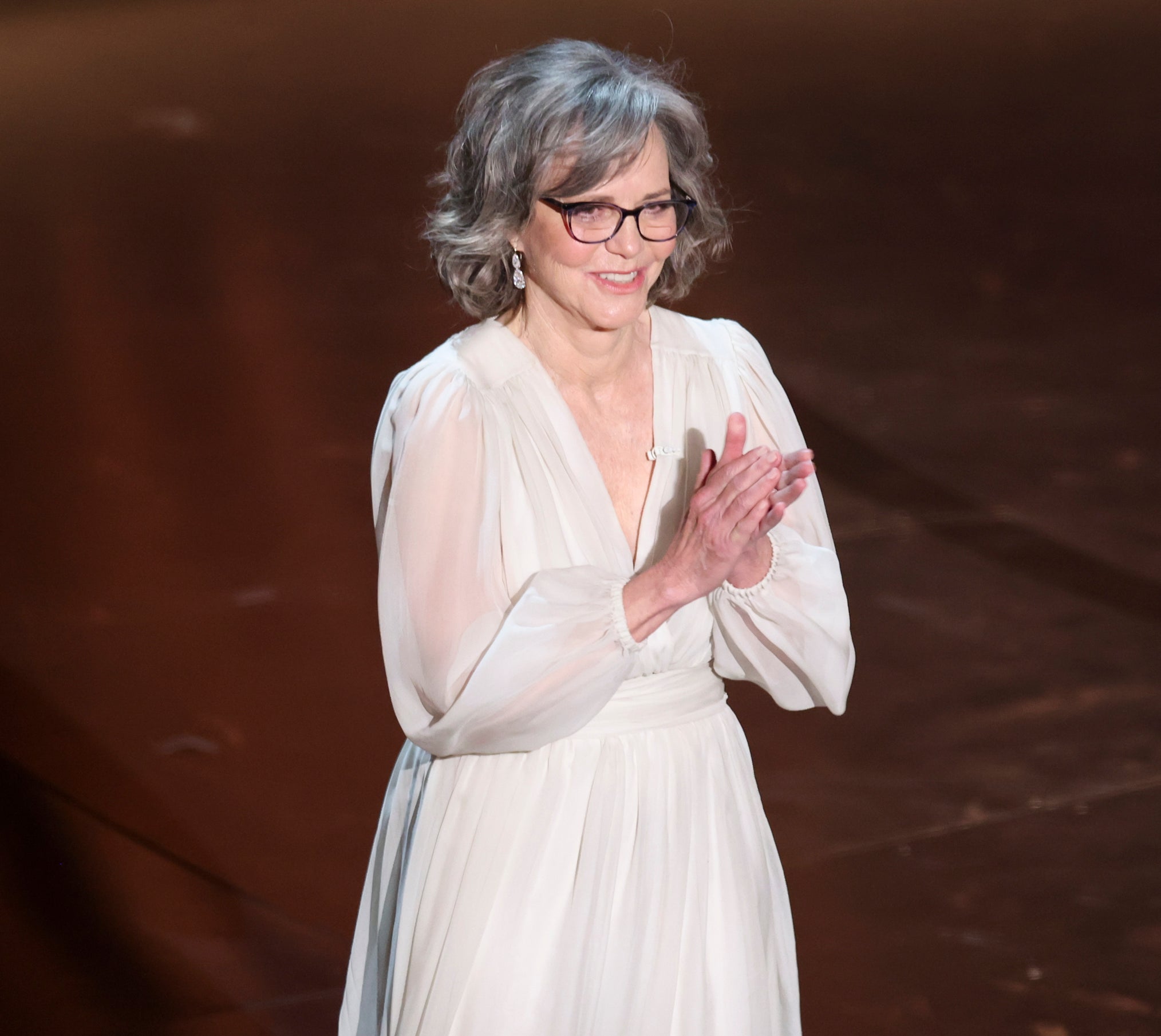 Sally Field stands clapping, wearing a v-neck dress with billowing sleeves