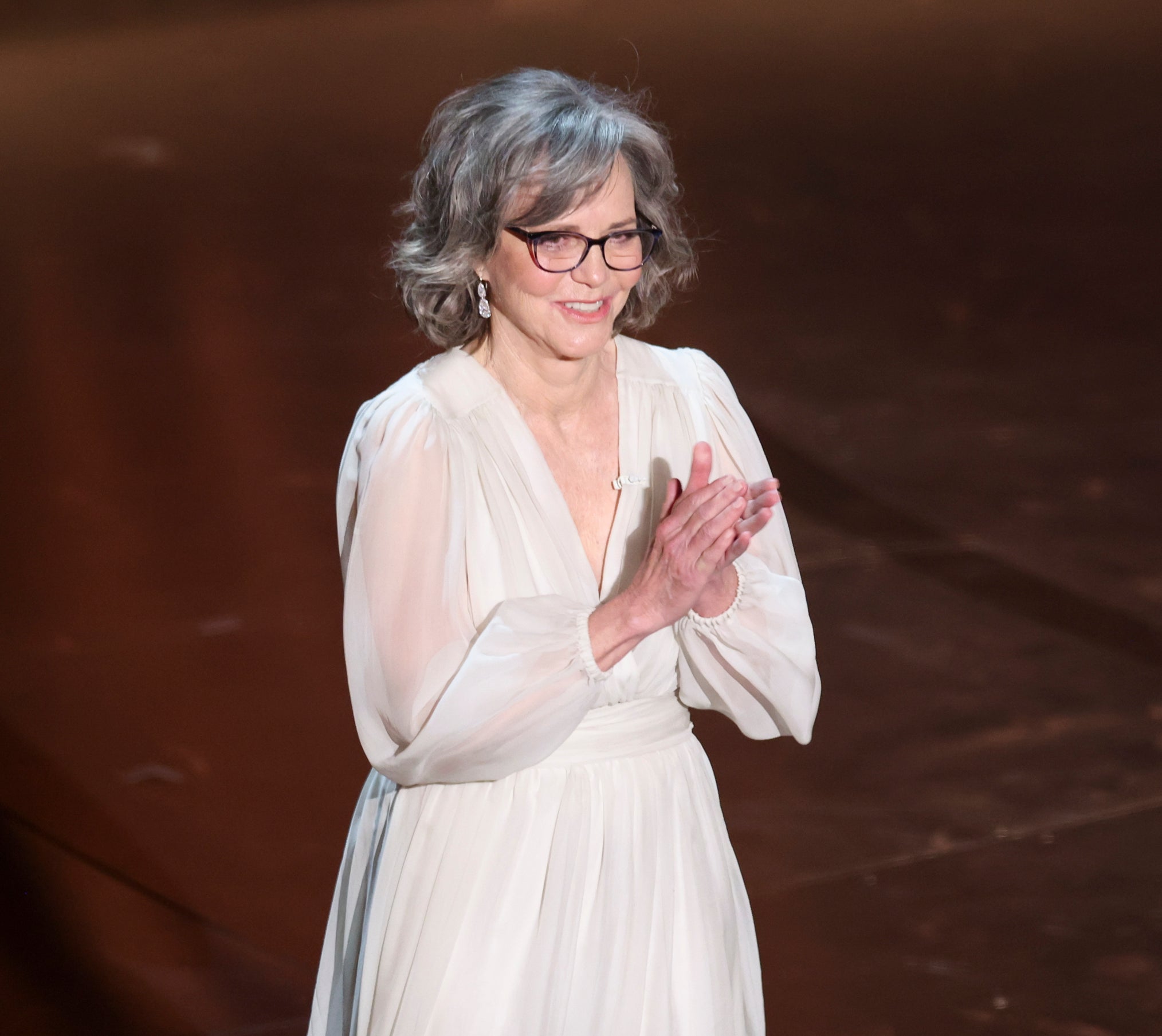 Sally Field stands clapping, wearing a v-neck dress with billowing sleeves