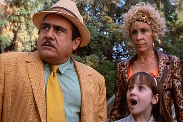 Danny DeVito, Rhea Perlman, and Mara Wilson stand together in a scene from &#x27;Matilda.&#x27; Danny wears a checkered suit, Rhea in a leopard print