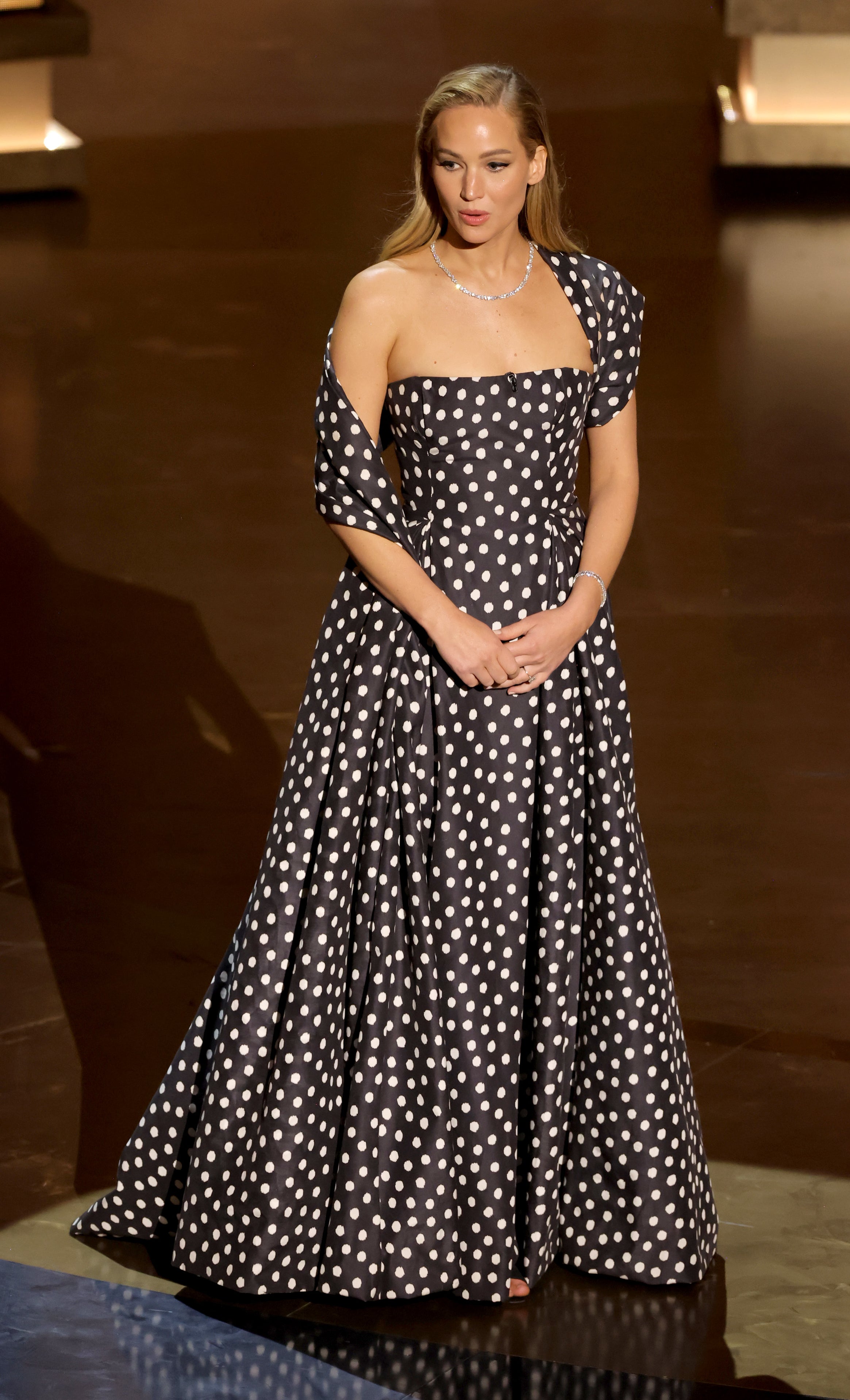 Jennifer in a polka dot off-shoulder gown and pearl necklace standing on stage