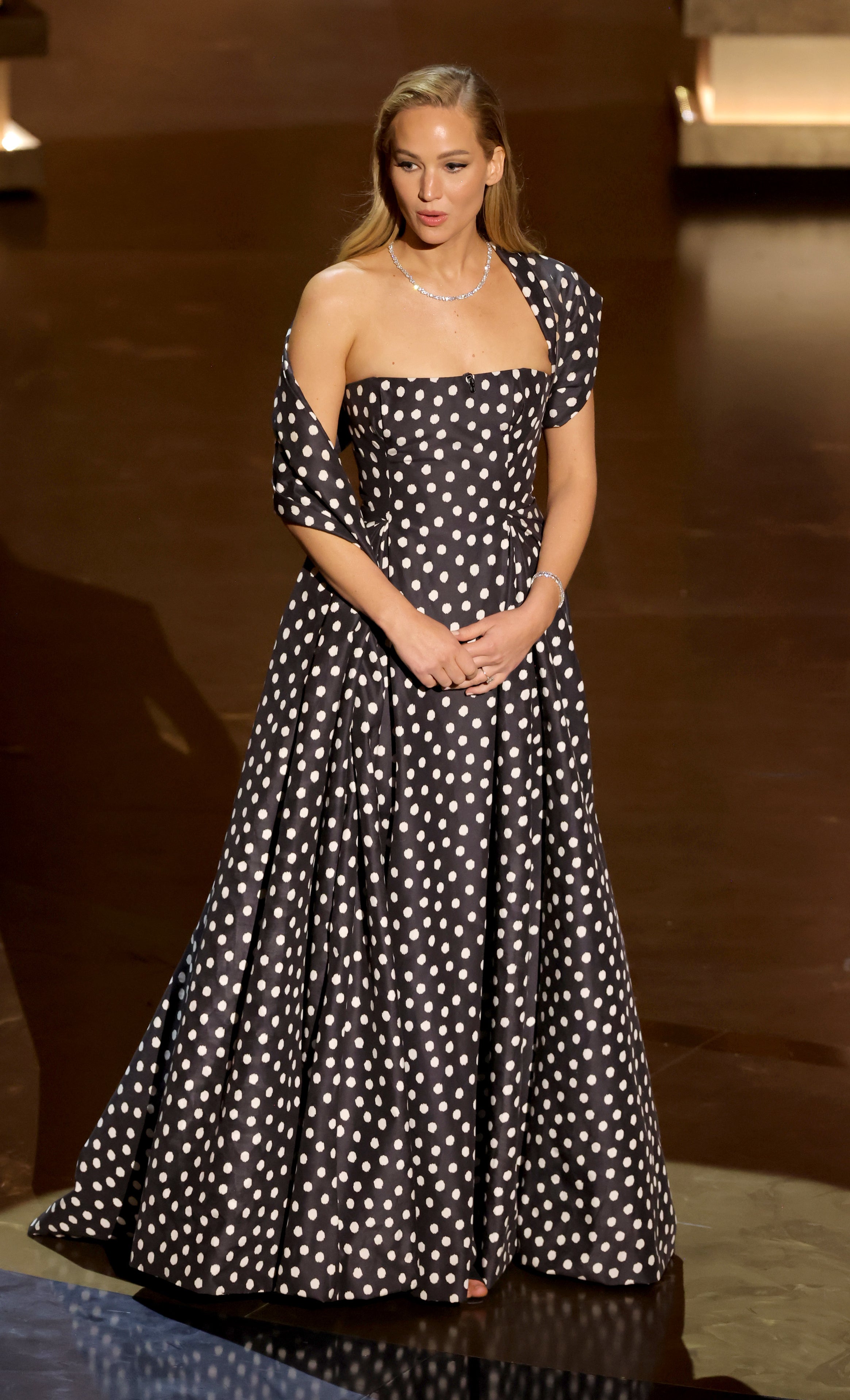 Jennifer in a polka dot off-shoulder gown and pearl necklace standing on stage