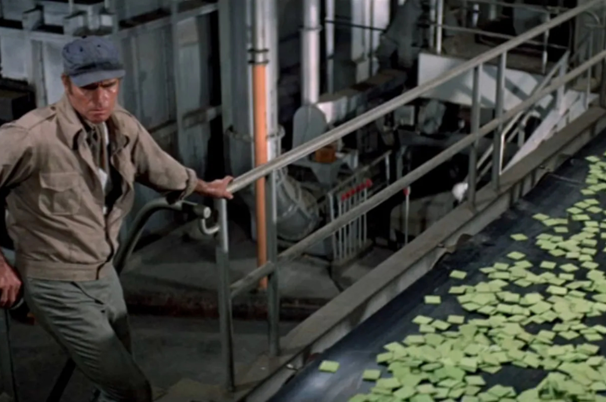 Man in a cap leaning over railing looking at papers on a conveyor belt in a factory