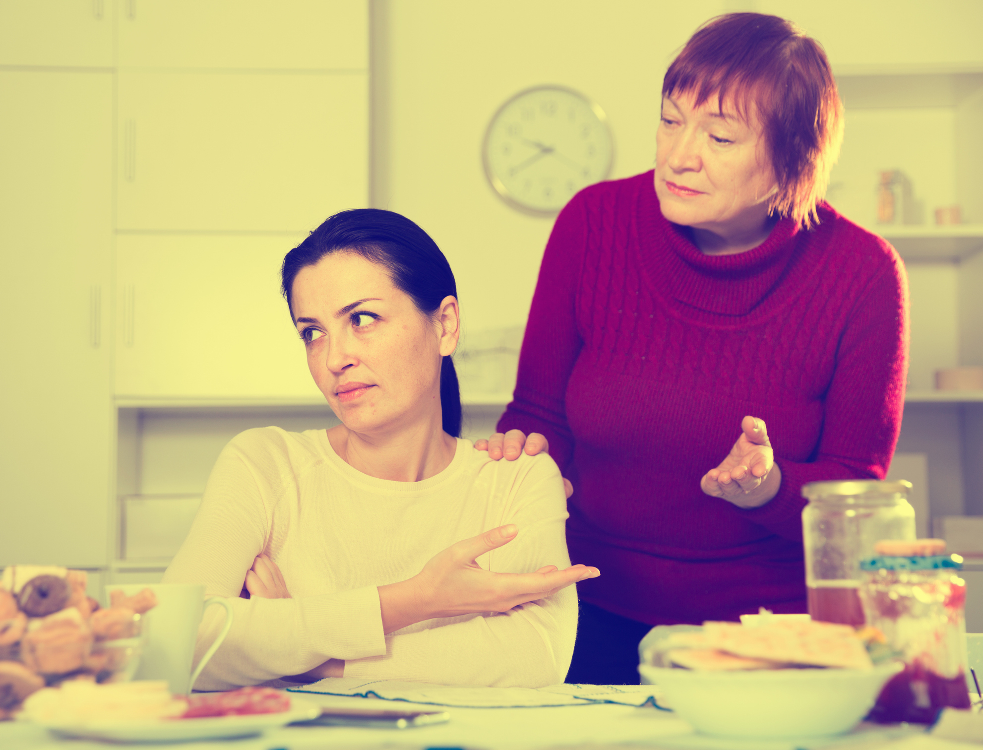 An older woman talks to a younger woman who looks upset at a kitchen table, suggesting a parent and adult child in a discussion
