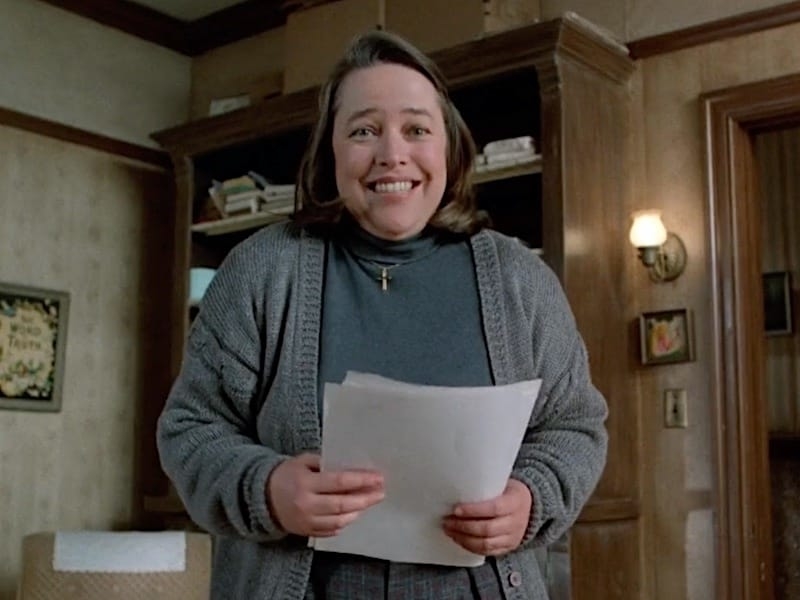 Character from &#x27;Misery&#x27; wearing a sweater and holding paper, smiling in a room