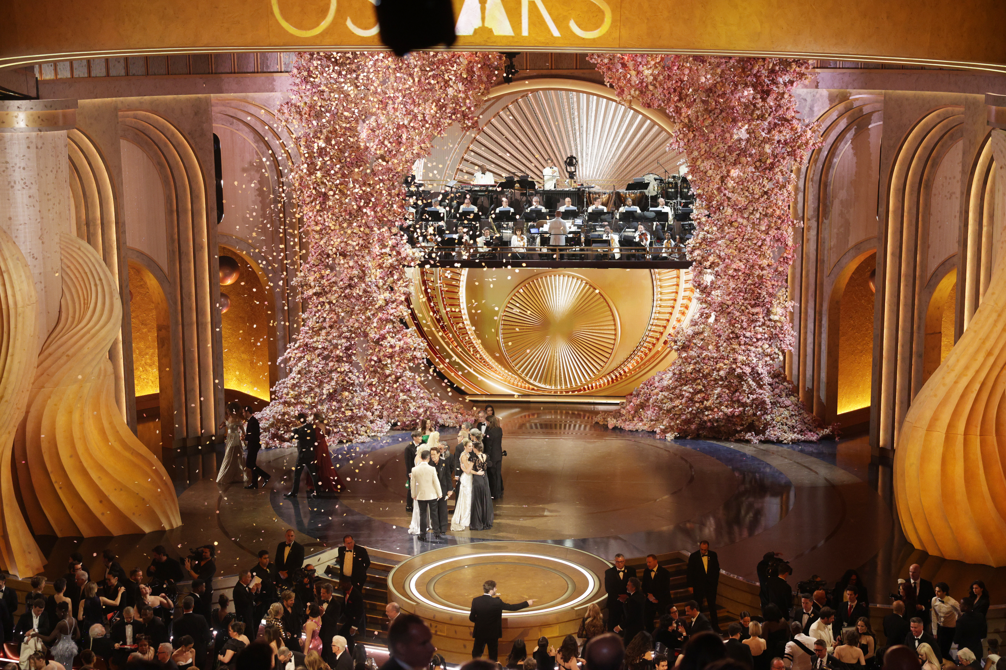 Elegant stage at Oscars with hosts speaking and orchestra above; audience in formal attire
