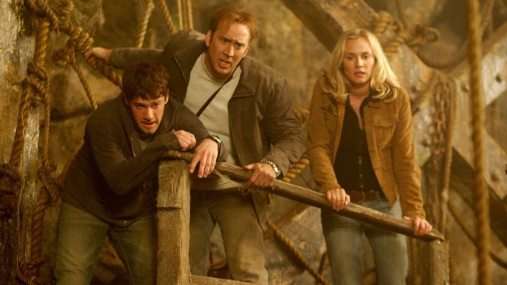 Three actors in an action scene, with intense expressions, leaning on a wooden structure