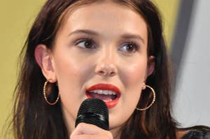 Millie Bobby Brown speaking into a microphone, wearing hoop earrings and a decorated top