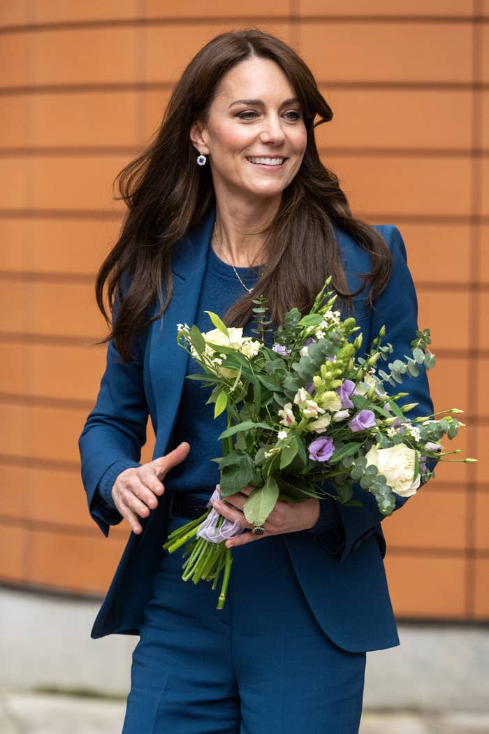 Kate Middleton smiling, wearing a blue blazer, holding a bouquet. She&#x27;s outside, near a brick wall