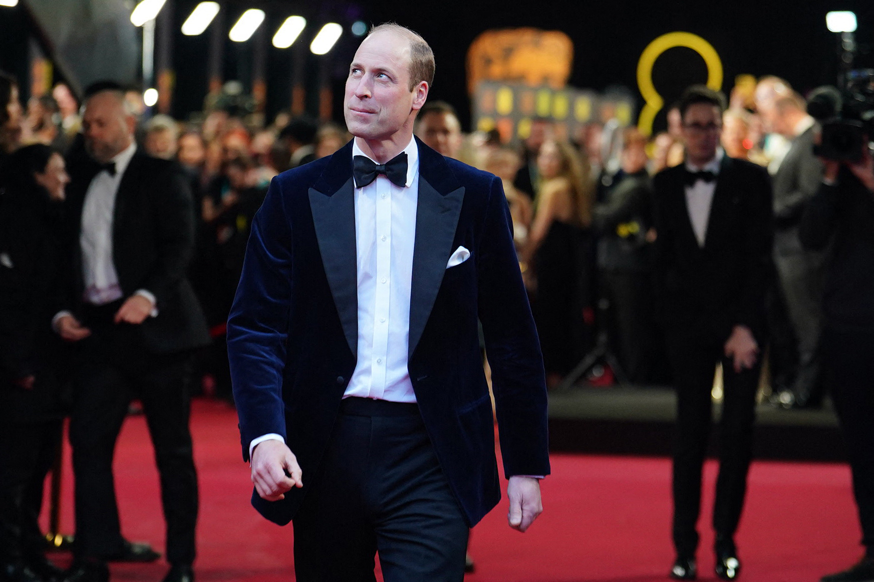 Prince William in a black tuxedo with a bow tie walks on a red carpet