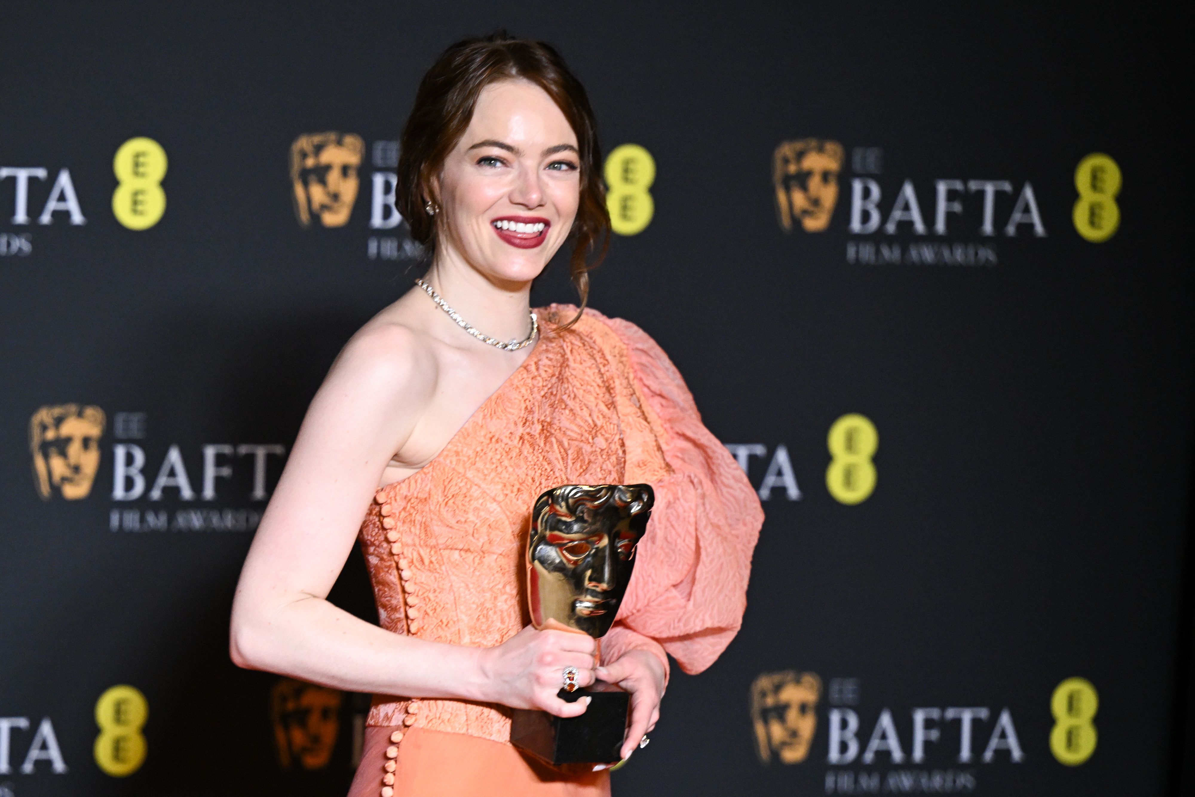 Emma Stone posing with her BAFTA award while wearing a one-shoulder dress