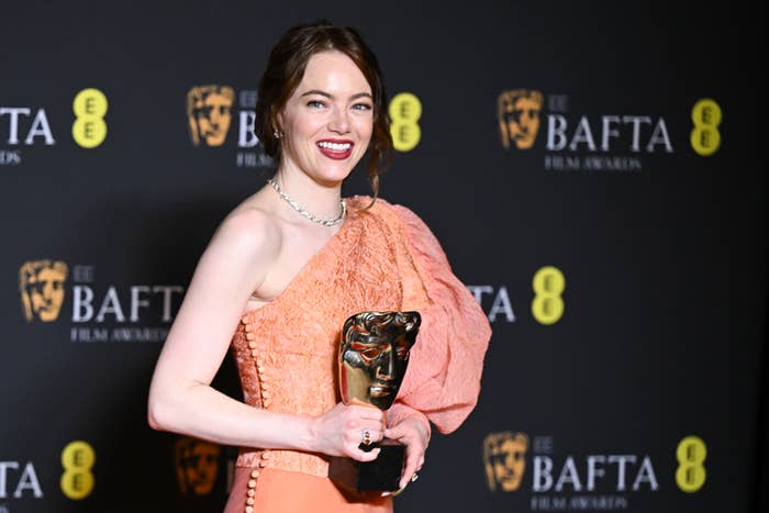 Emma Stone posing with a BAFTA award, wearing a strapless dress and a smile
