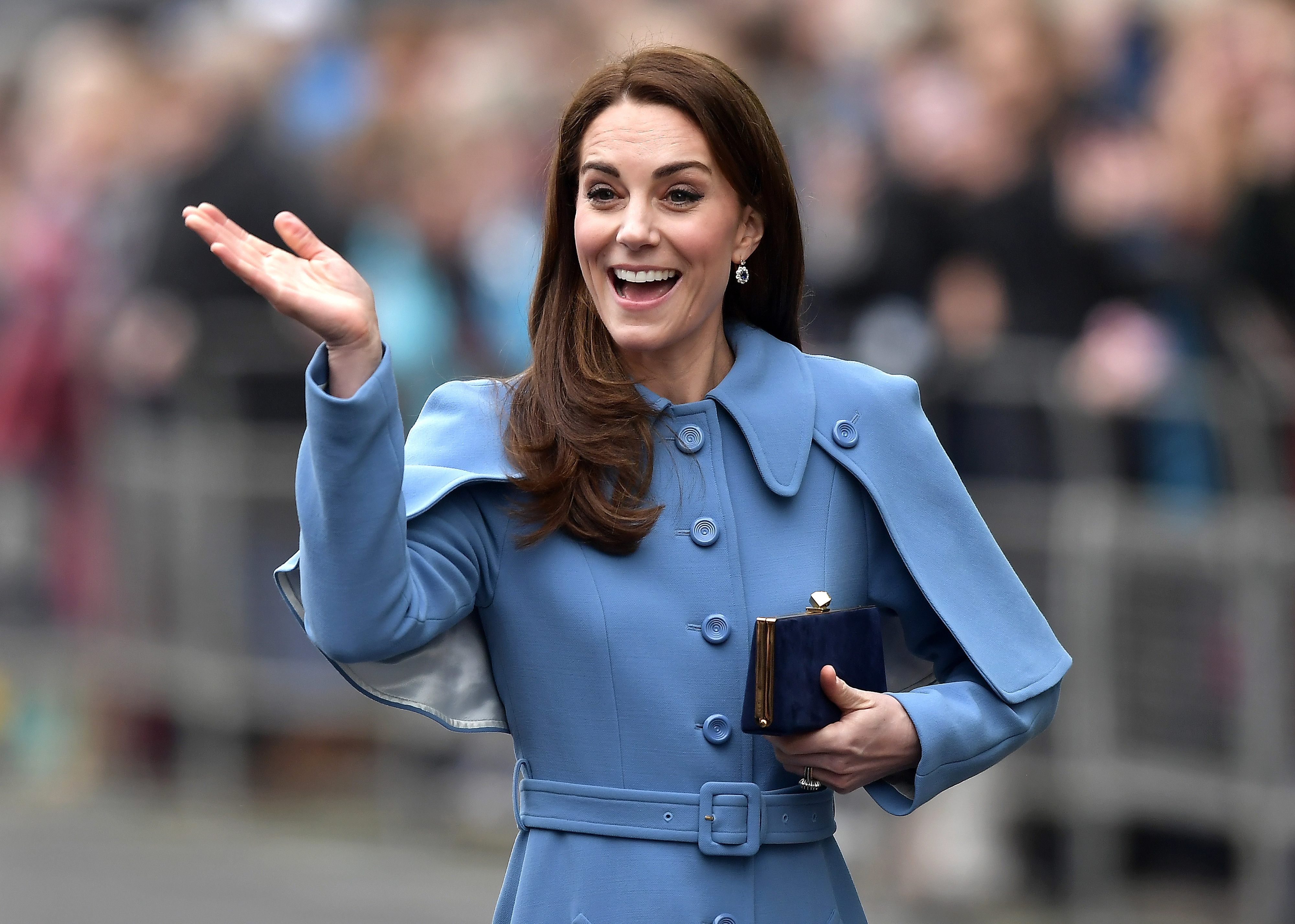Kate Middleton in blue coat waves with her right hand, smiling with onlookers in the background