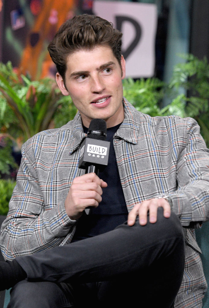 Gregg Sulkin wearing a plaid jacket, holding a microphone, seated during an interview