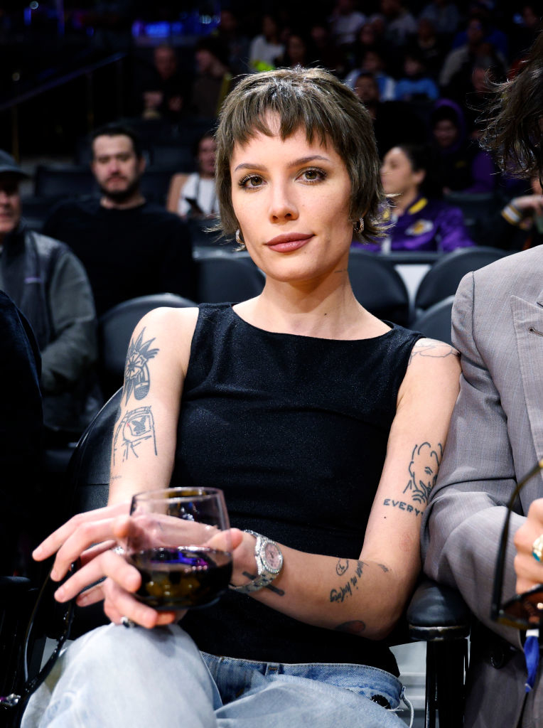 halsey seated at a sports event, wearing a sleeveless top and jeans with a glass of wine in hand