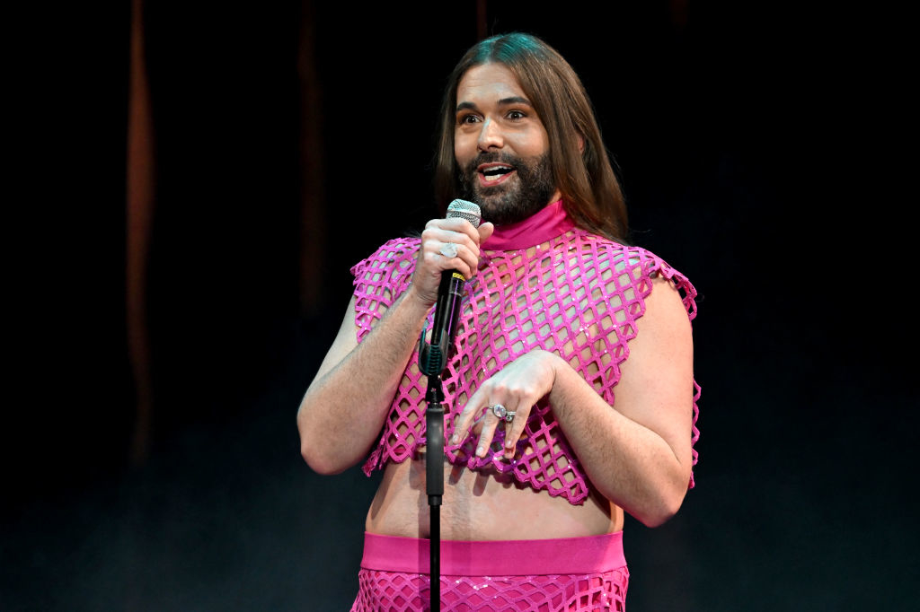 Jonathan Van Ness wearing a fishnet top and pants, speaking into a microphone on stage