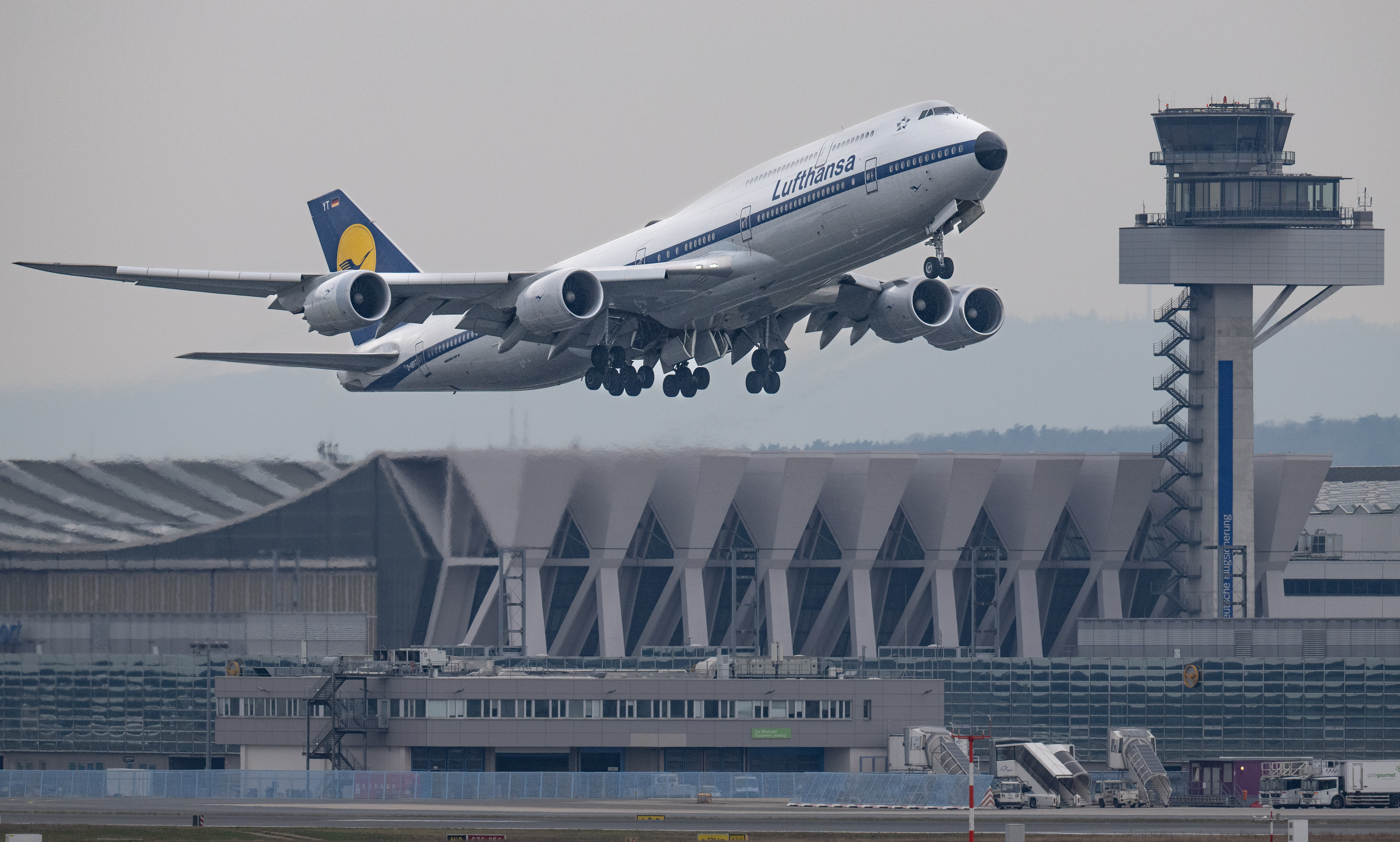 A Lufthansa airplane takes off from an airport, with the terminal visible in the background